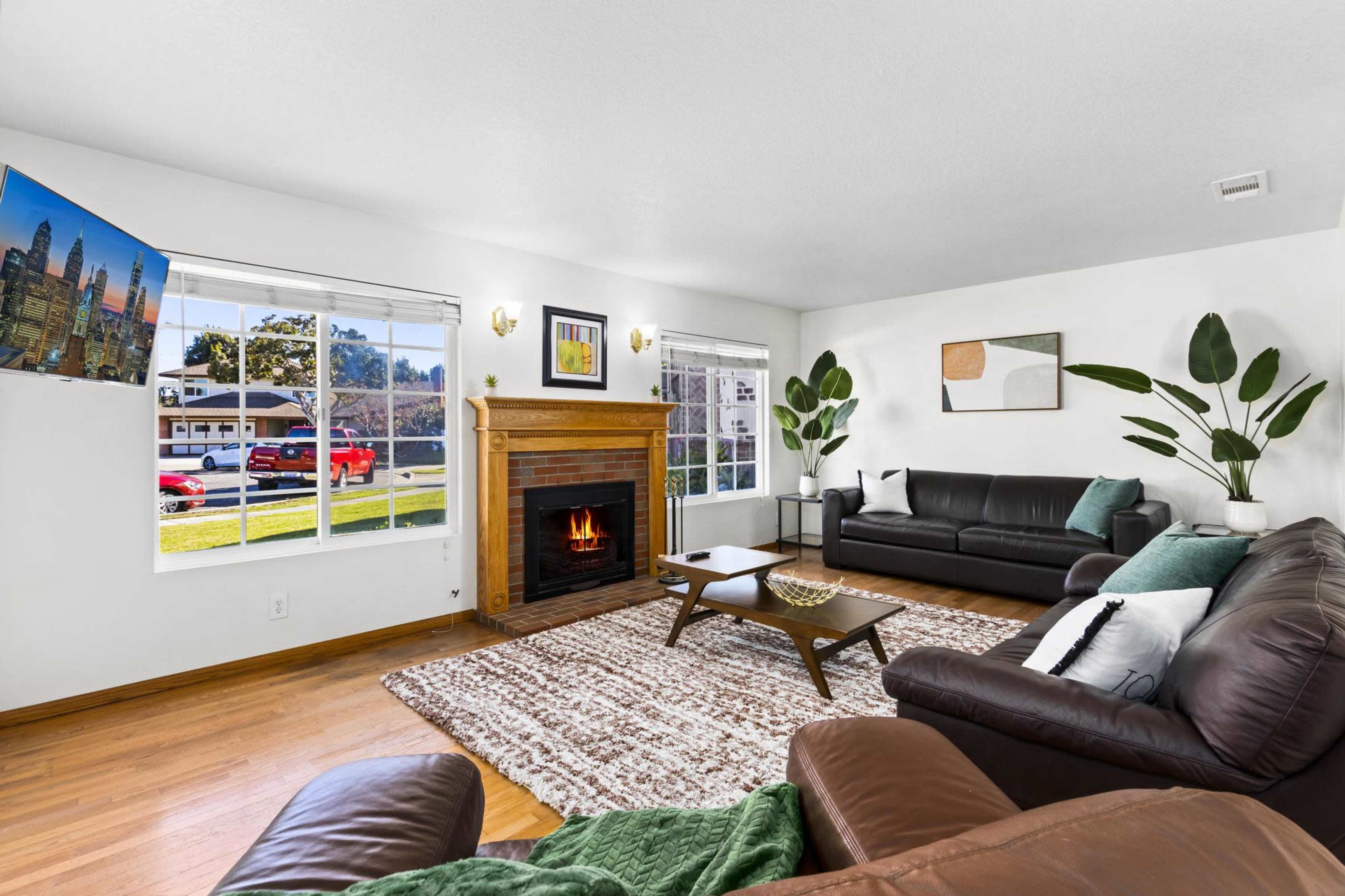 A living room features two black sofas, a wooden fireplace with a fire burning, a large window facing the street, and a wall-mounted television.