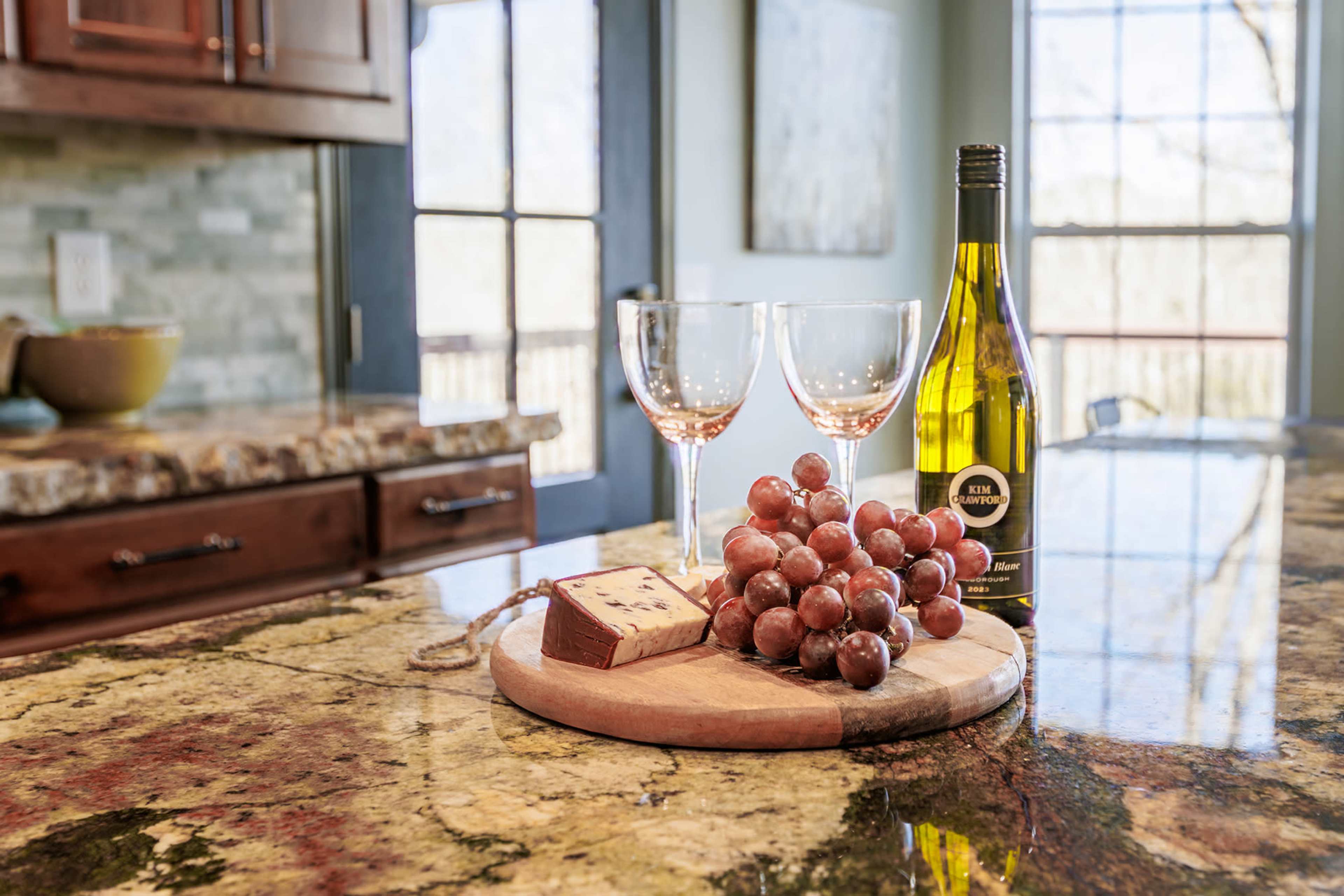 A platter with cheese, grapes, and a bottle of wine sits on a granite countertop next to two wine glasses in a well-lit kitchen.