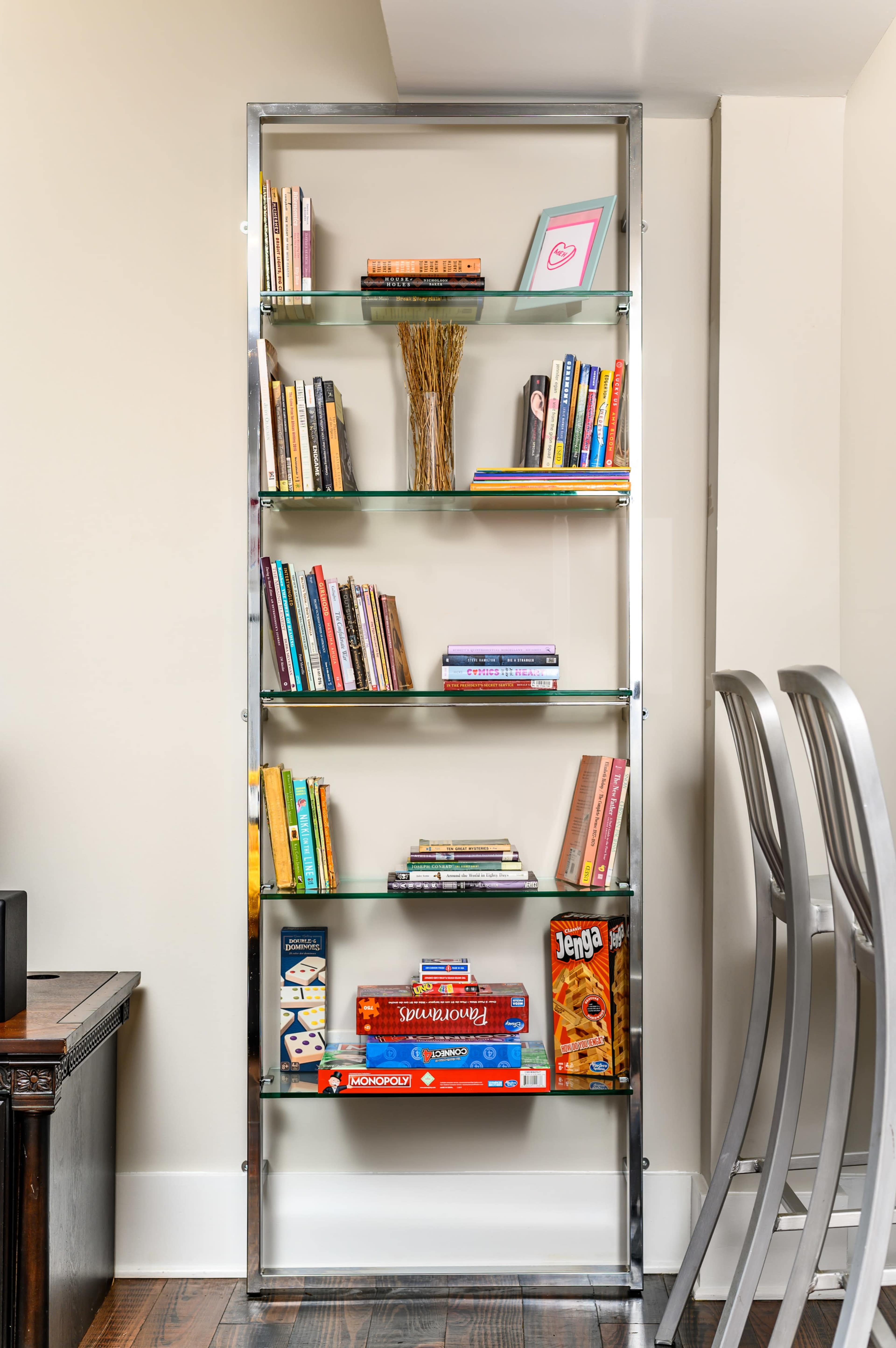 A metal and glass bookshelf displays an assortment of books and board games, including Jenga, in a well-lit room.