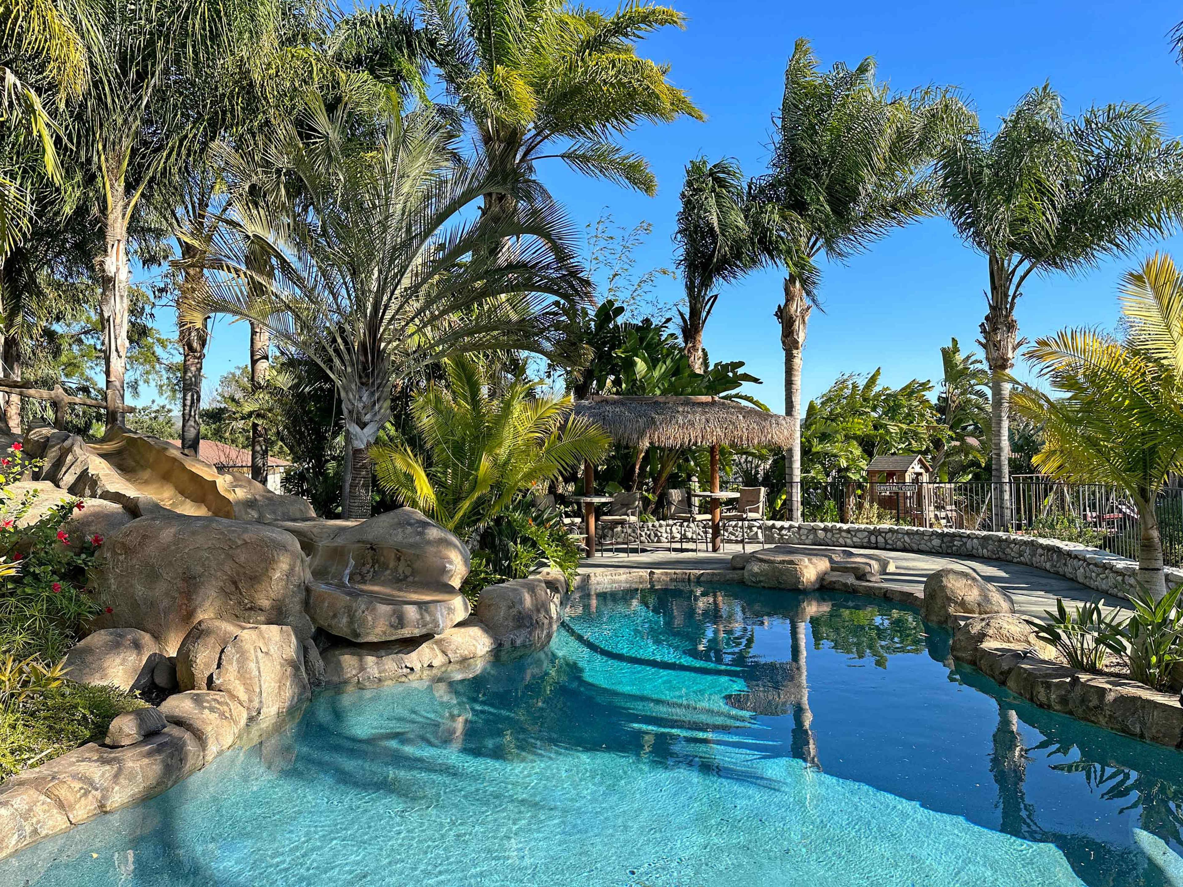 The image features a vibrant pool area surrounded by lush palm trees and tropical plants, with a rock formation and a thatched gazebo nearby.