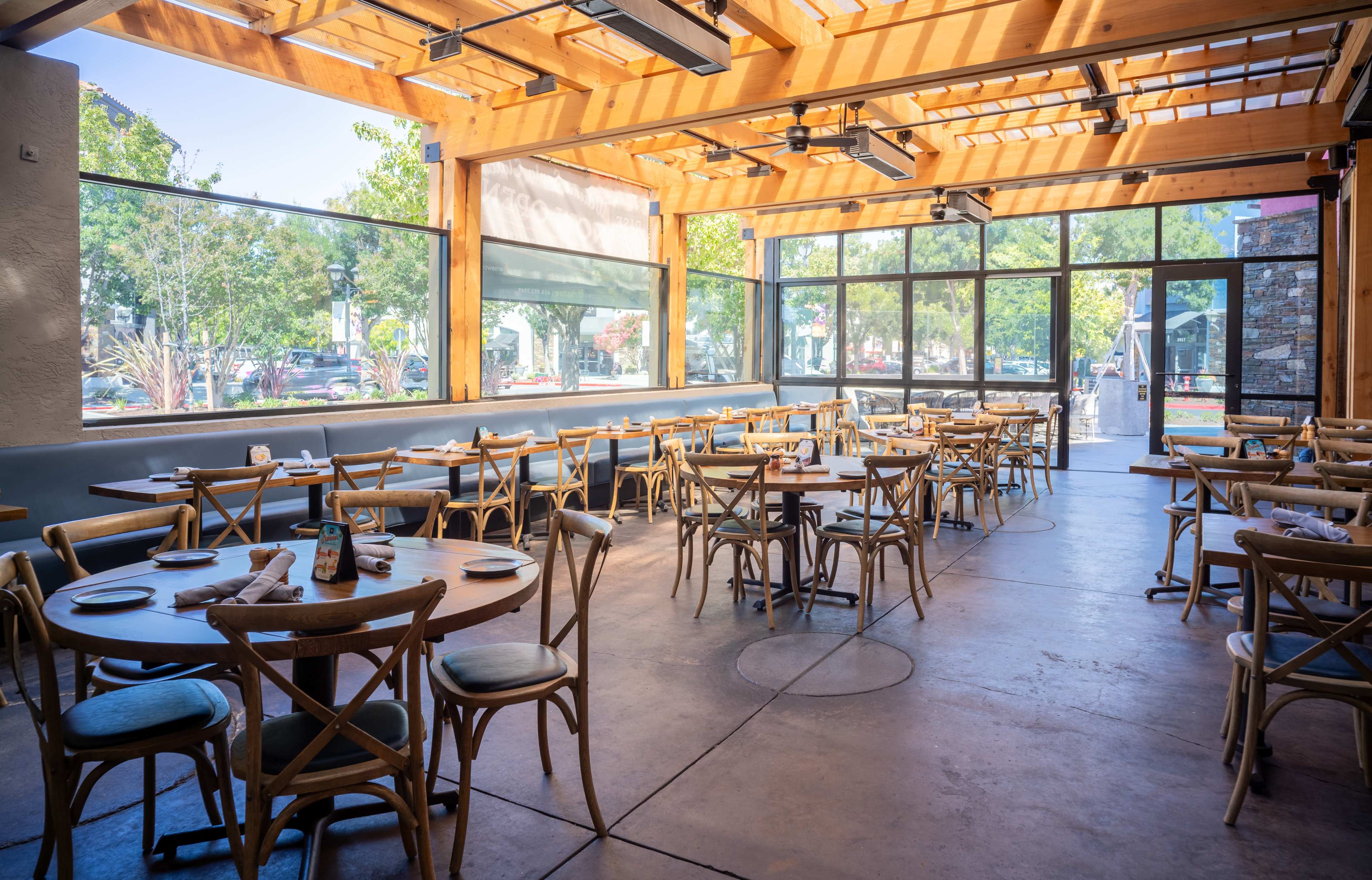The image shows a bright and spacious restaurant interior with wooden tables and chairs arranged neatly, large windows letting in natural light, and a view of the outside.
