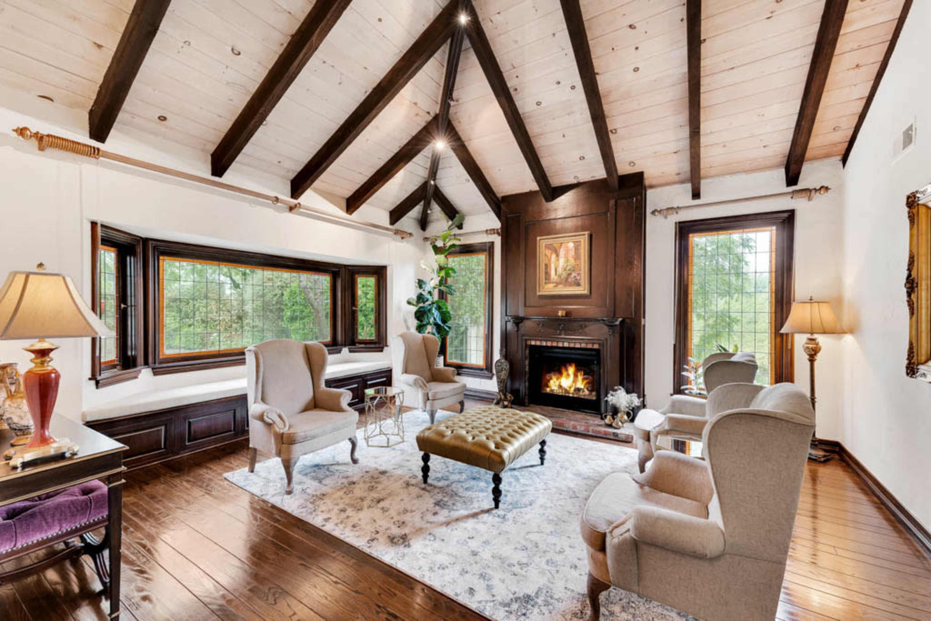 The room features a cozy sitting area with four chairs arranged around a central coffee table, a fireplace set into a wooden wall, and large windows that provide views of the greenery outside.