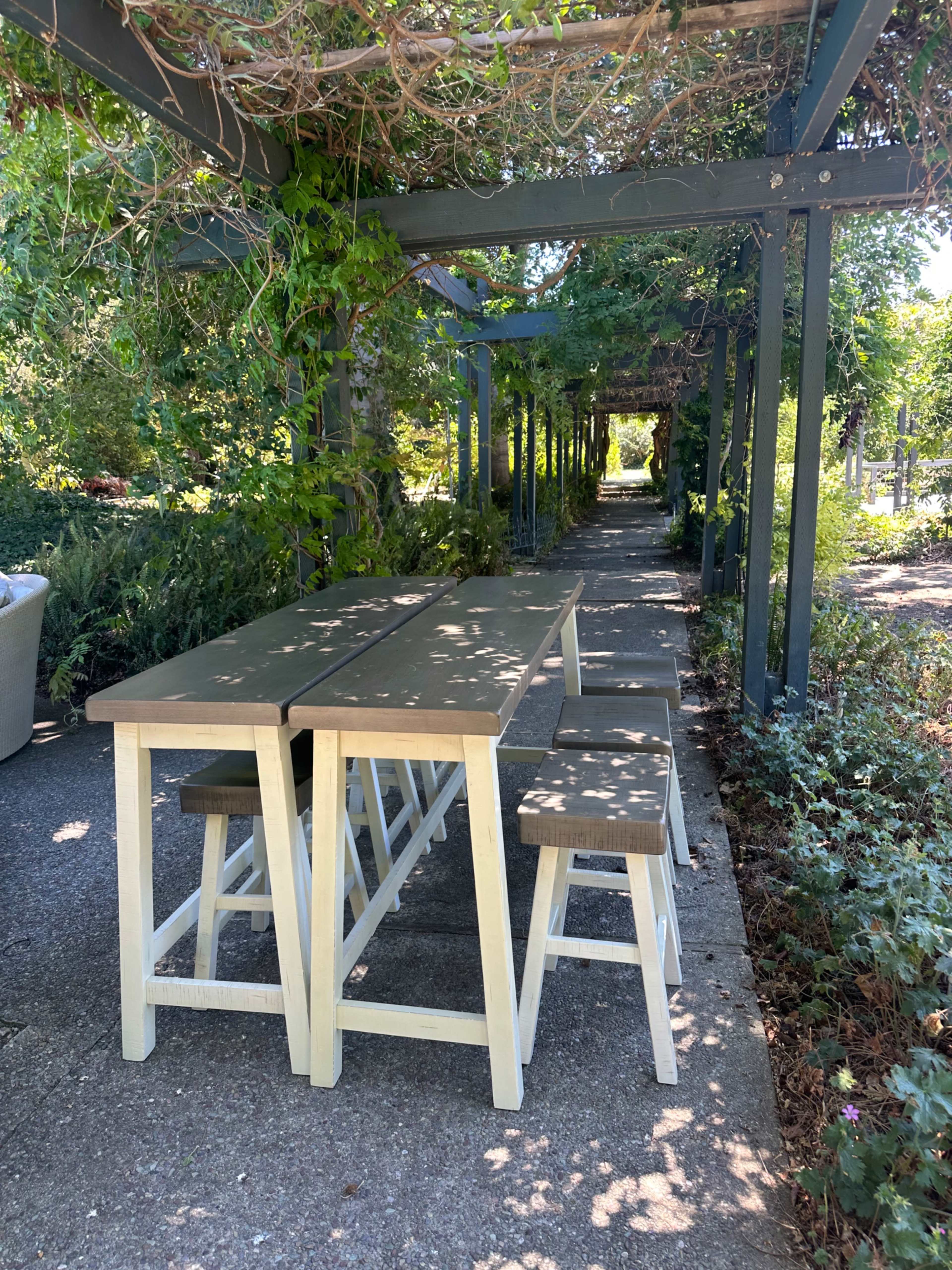 A long table with stools is set under a leafy pergola along a paved walkway surrounded by greenery.