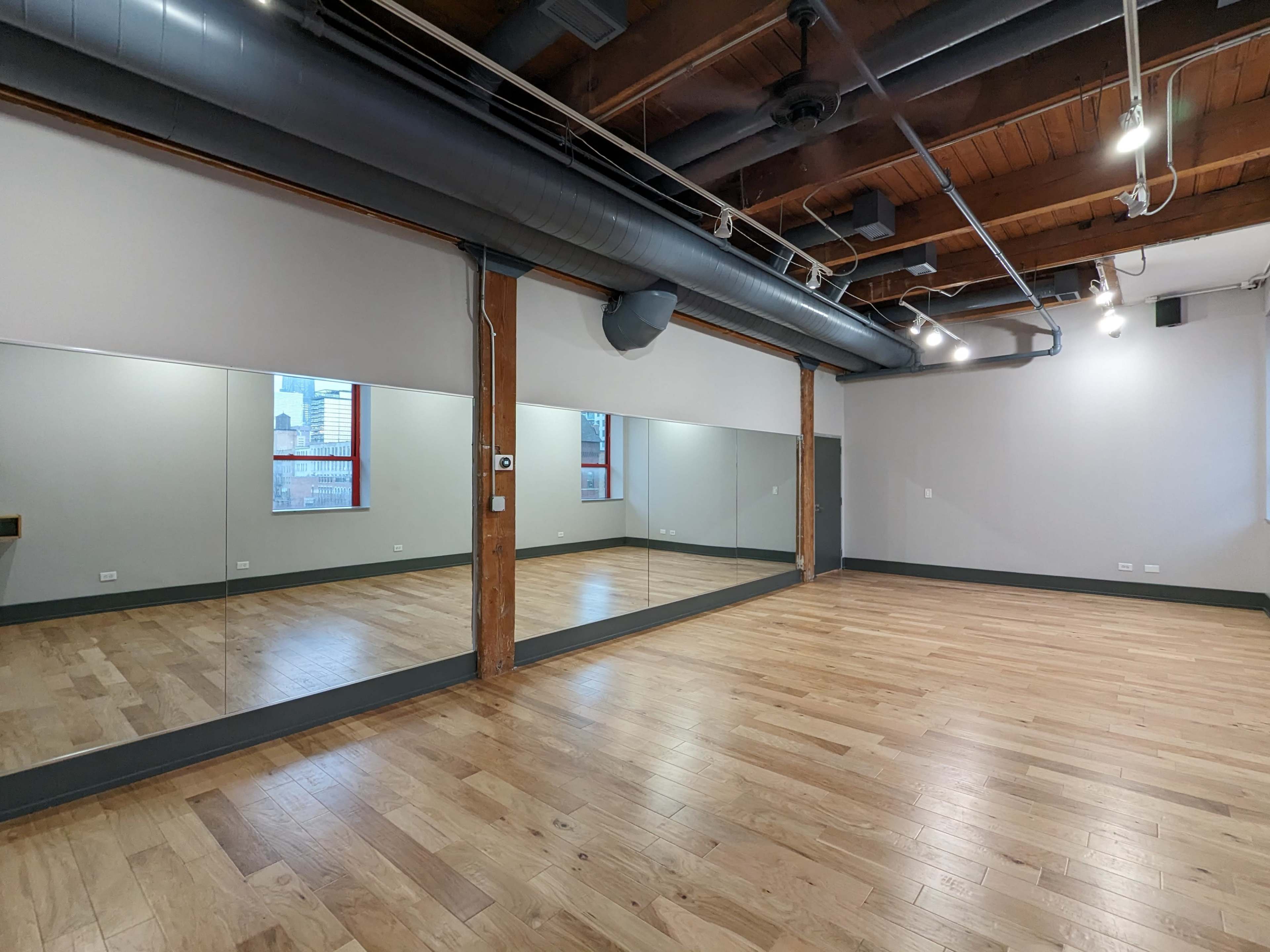 A spacious studio with wooden floors, large mirrors on one wall, and exposed wooden beams and pipes on the ceiling.