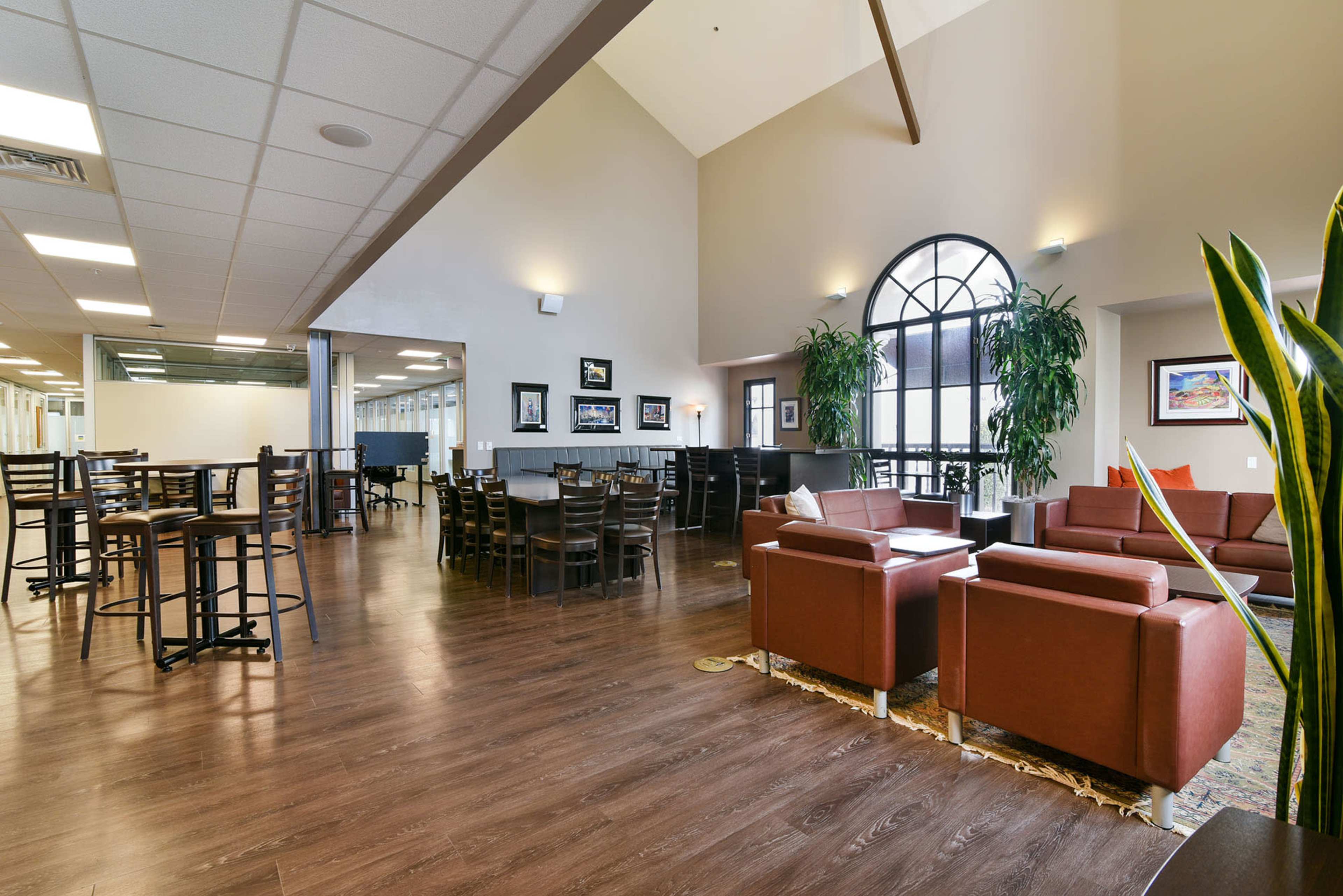 A spacious indoor dining area with wooden flooring, tables, chairs, and large windows, complemented by green plants and comfortable seating arrangements.