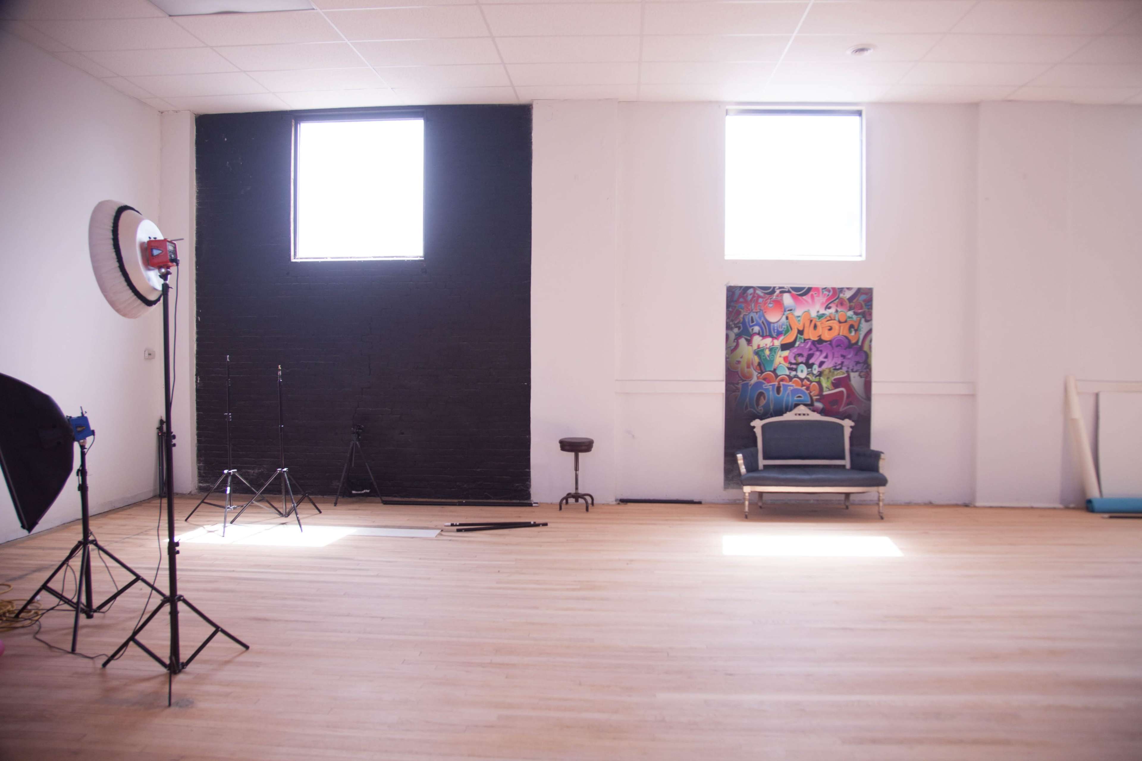 A spacious room with hardwood floors, two windows, a black wall, a colorful graffiti mural, and a photography setup including a light stand and a stool.