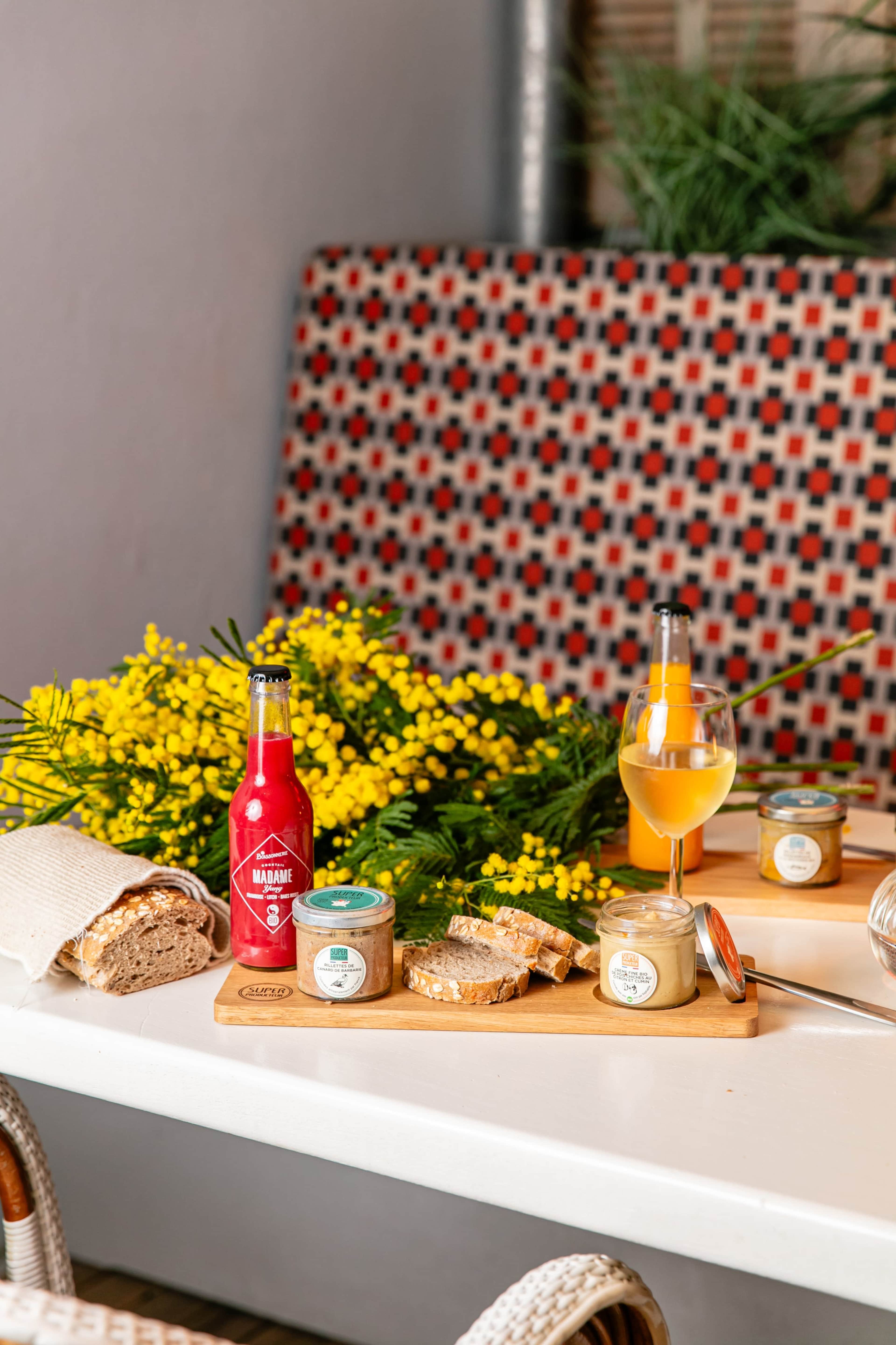 A wooden platter displays jars of spreads, slices of bread, and a beverage beside a vibrant arrangement of yellow flowers on a table.