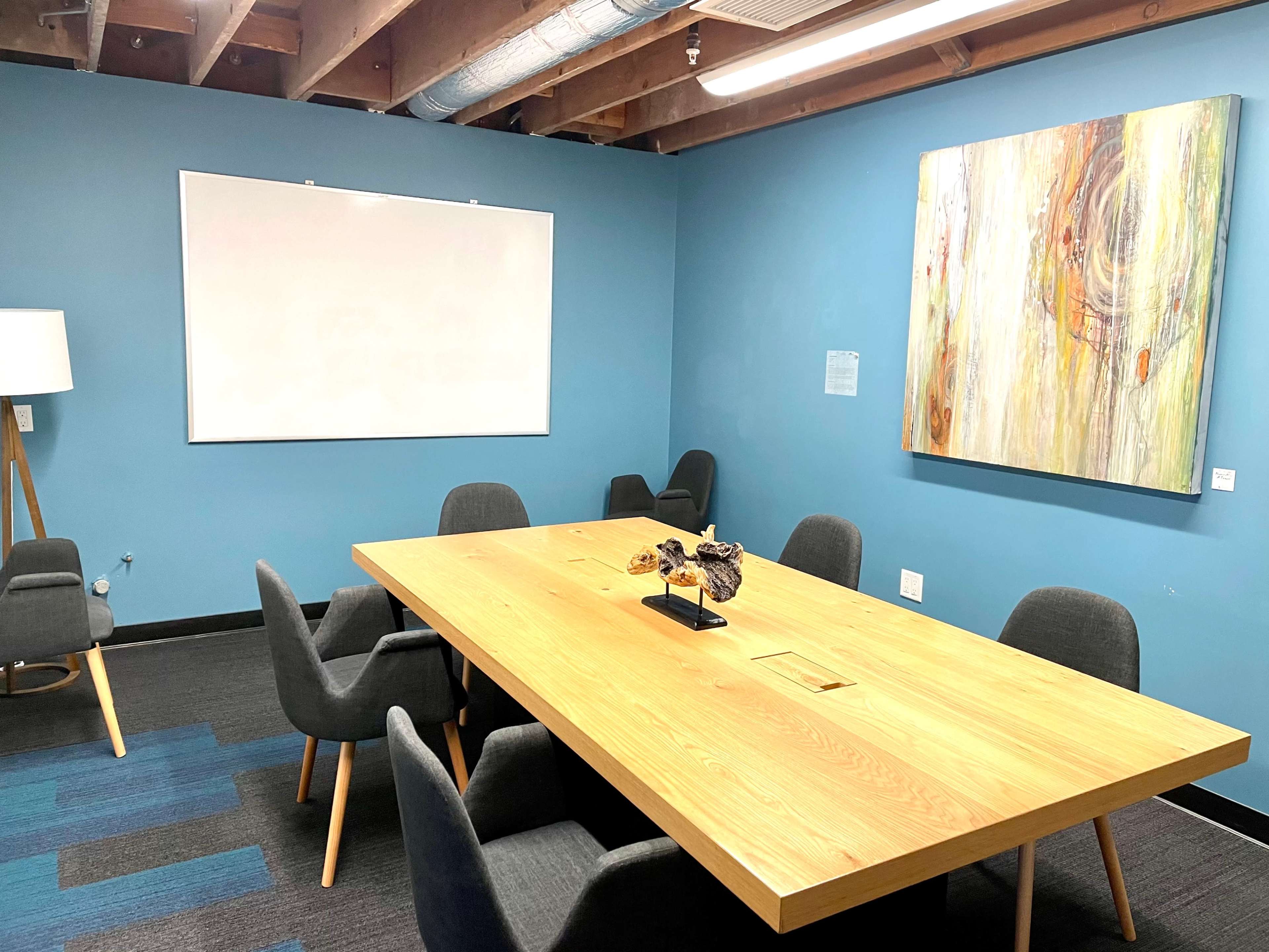 A modern conference room with a large wooden table surrounded by gray chairs, a whiteboard on the wall, and a colorful abstract painting.