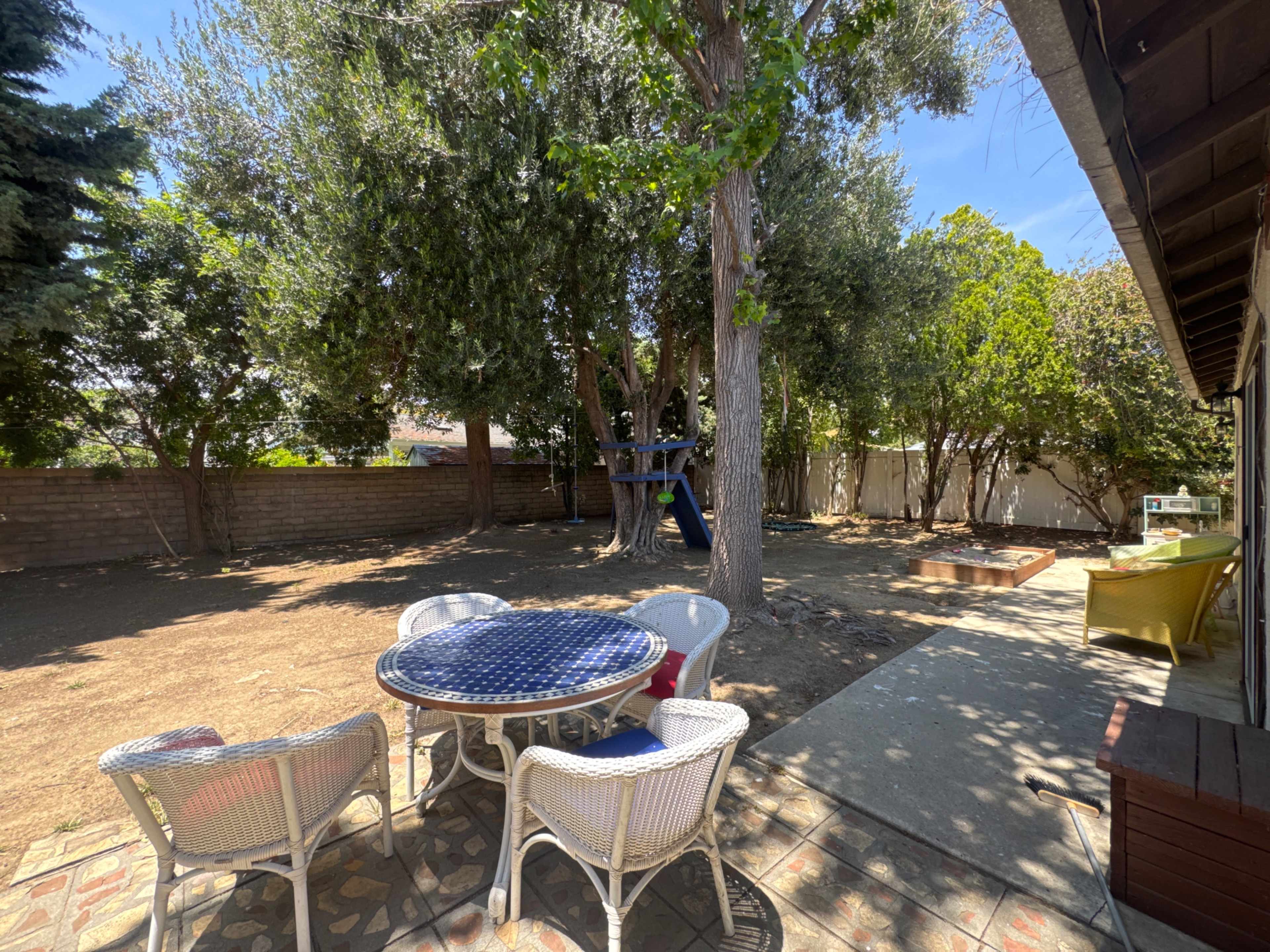 A backyard scene features a round table surrounded by white chairs, with a play structure visible in the background among trees and a patch of bare ground.