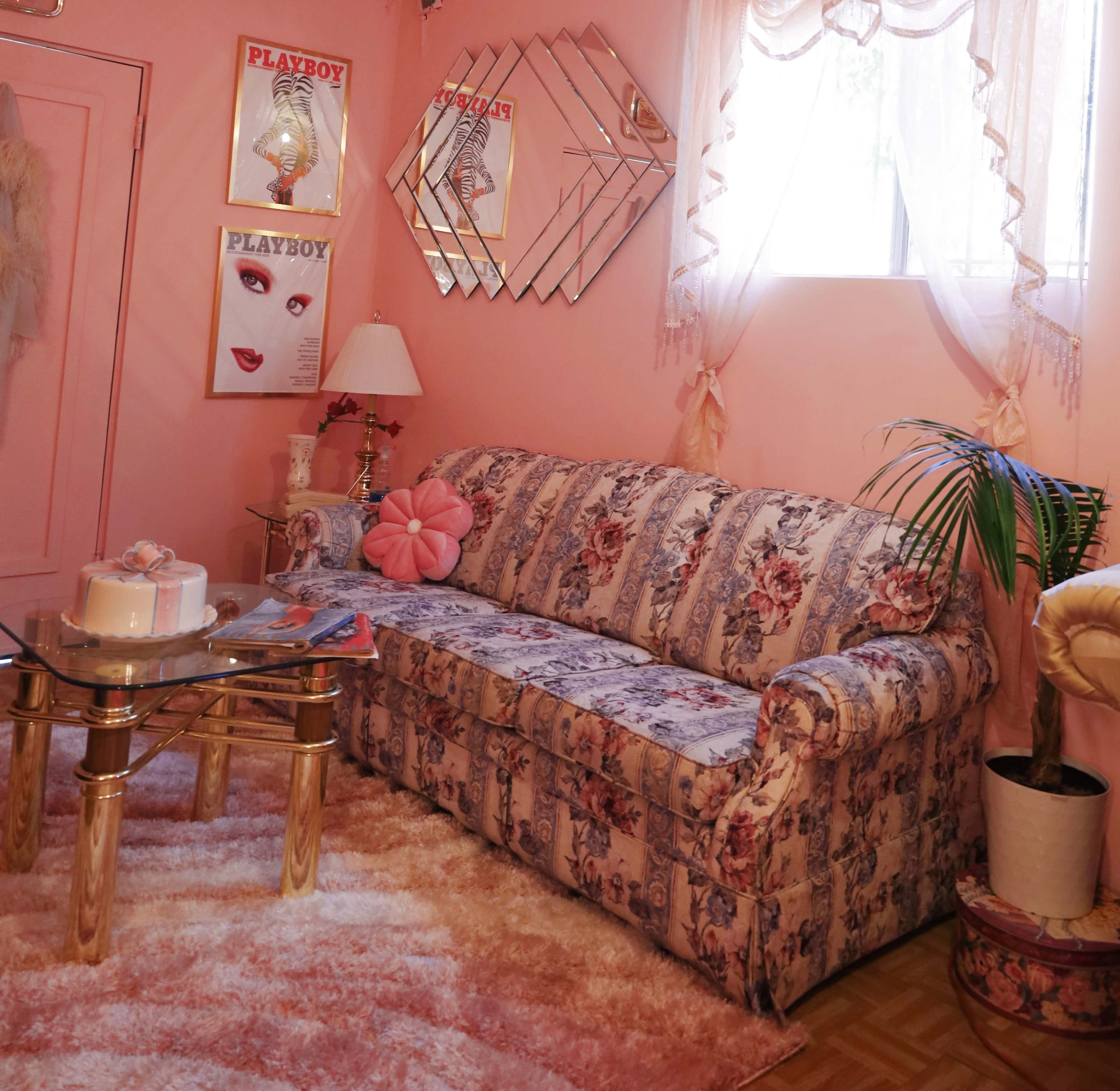 A pink-themed living room with a floral-patterned sofa, a glass coffee table, and decorative posters on the wall.