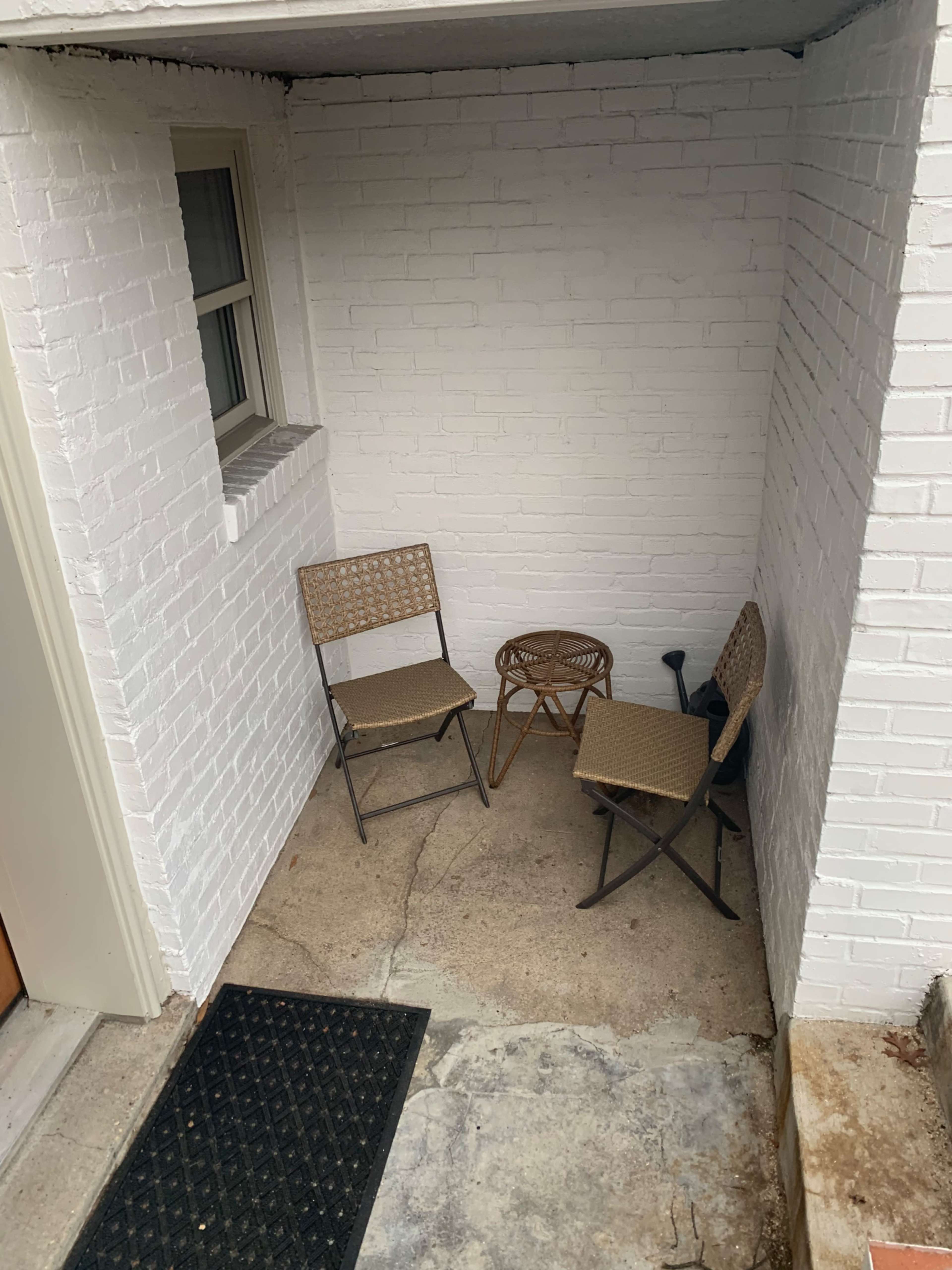 A small, corner seating area with two metal chairs and a round wicker table, situated against a white brick wall.