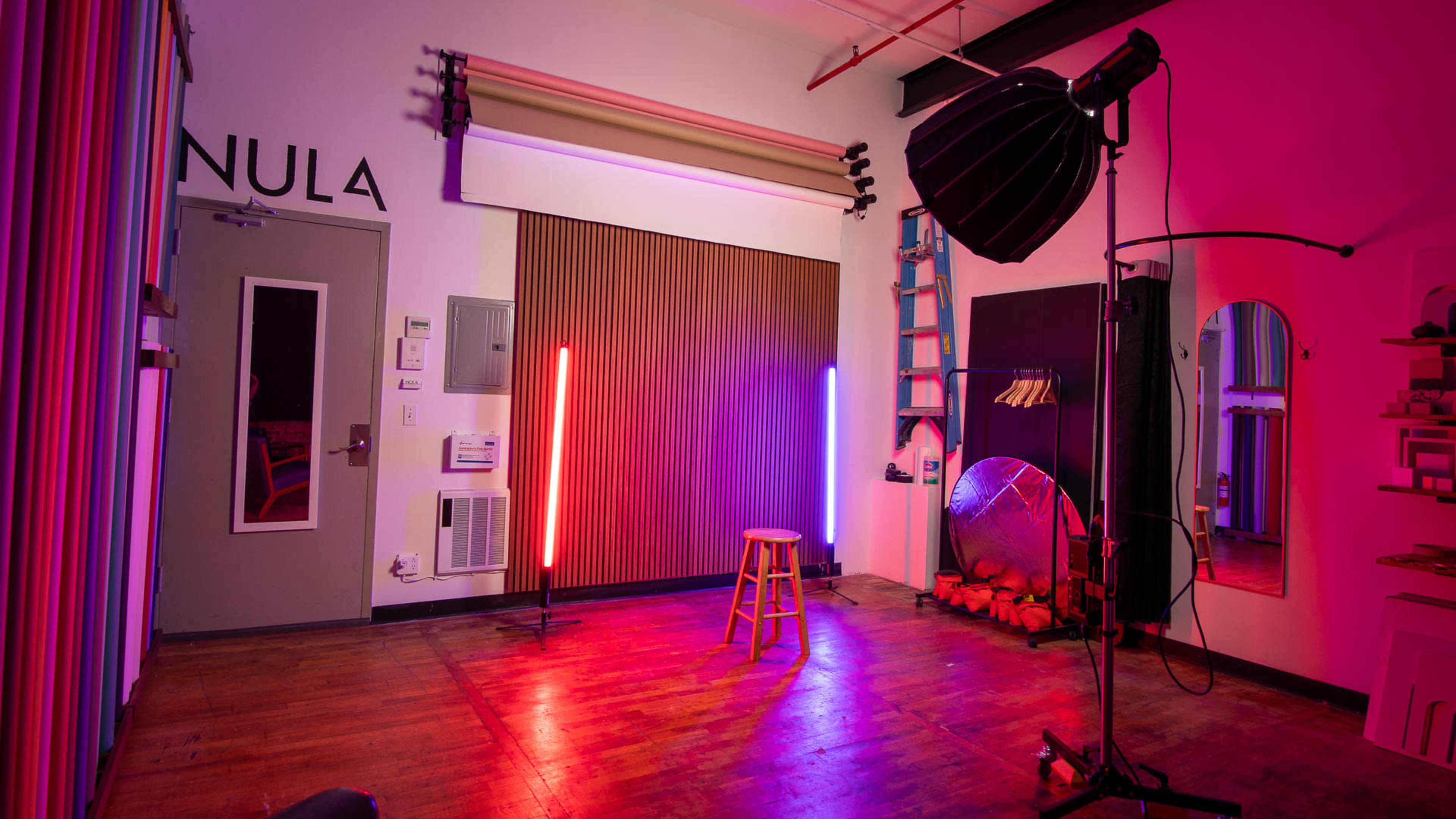 A photography studio features a stool in the center, flanked by neon lights, with a large softbox and reflective surfaces visible in the background.
