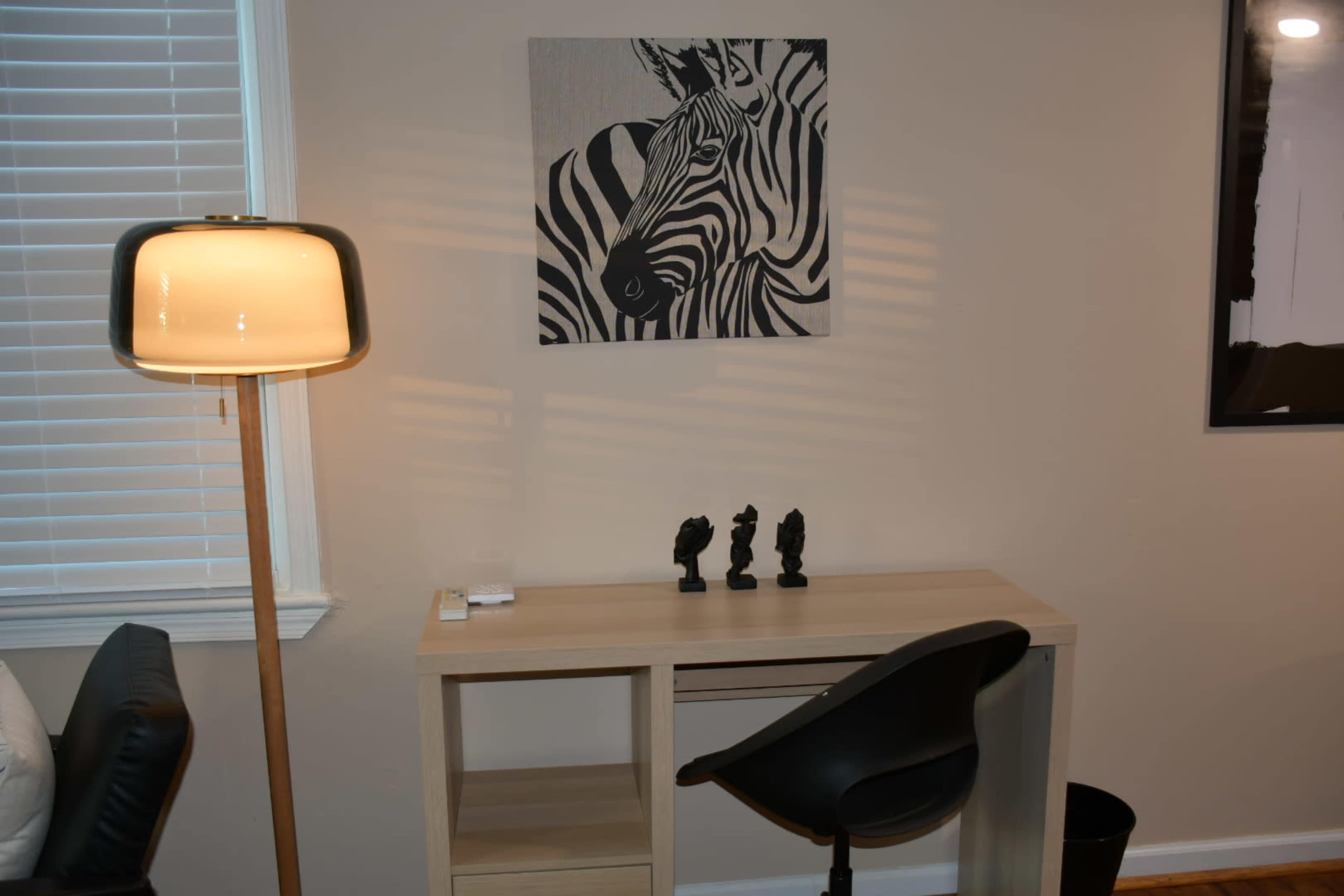 A minimalist workspace features a wooden desk with a chair, small decorative figures, a lamp, and a zebra print artwork on the wall.