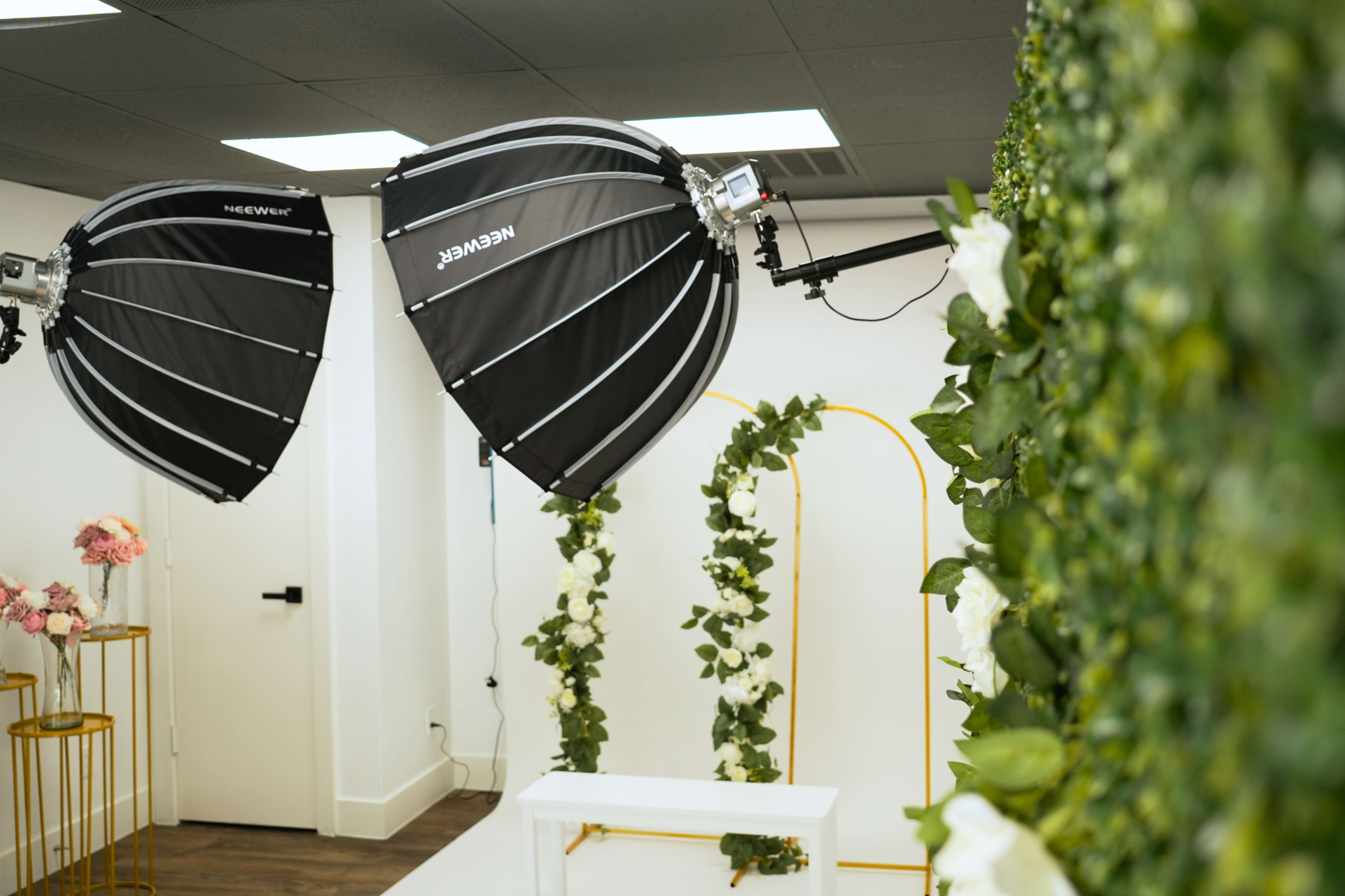 The image shows a photography studio setup featuring two large softbox lights positioned above a floral backdrop adorned with greenery and white flowers, alongside a decorative arch and a small white table.