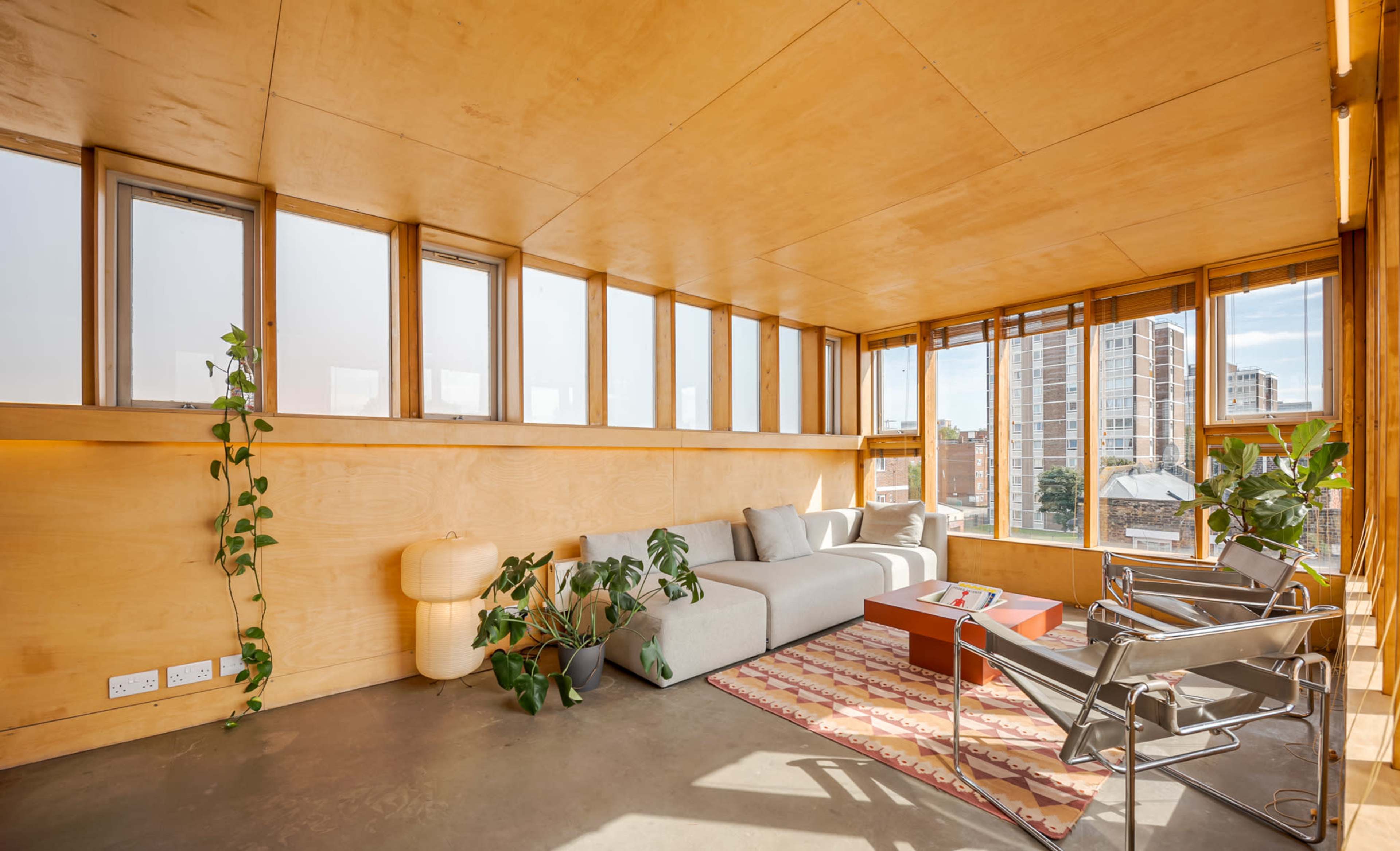 The image shows a modern interior living space with large windows, a light wood-paneled wall, a gray sofa, two metal chairs, and a small table, along with plants and a patterned rug on a concrete floor.