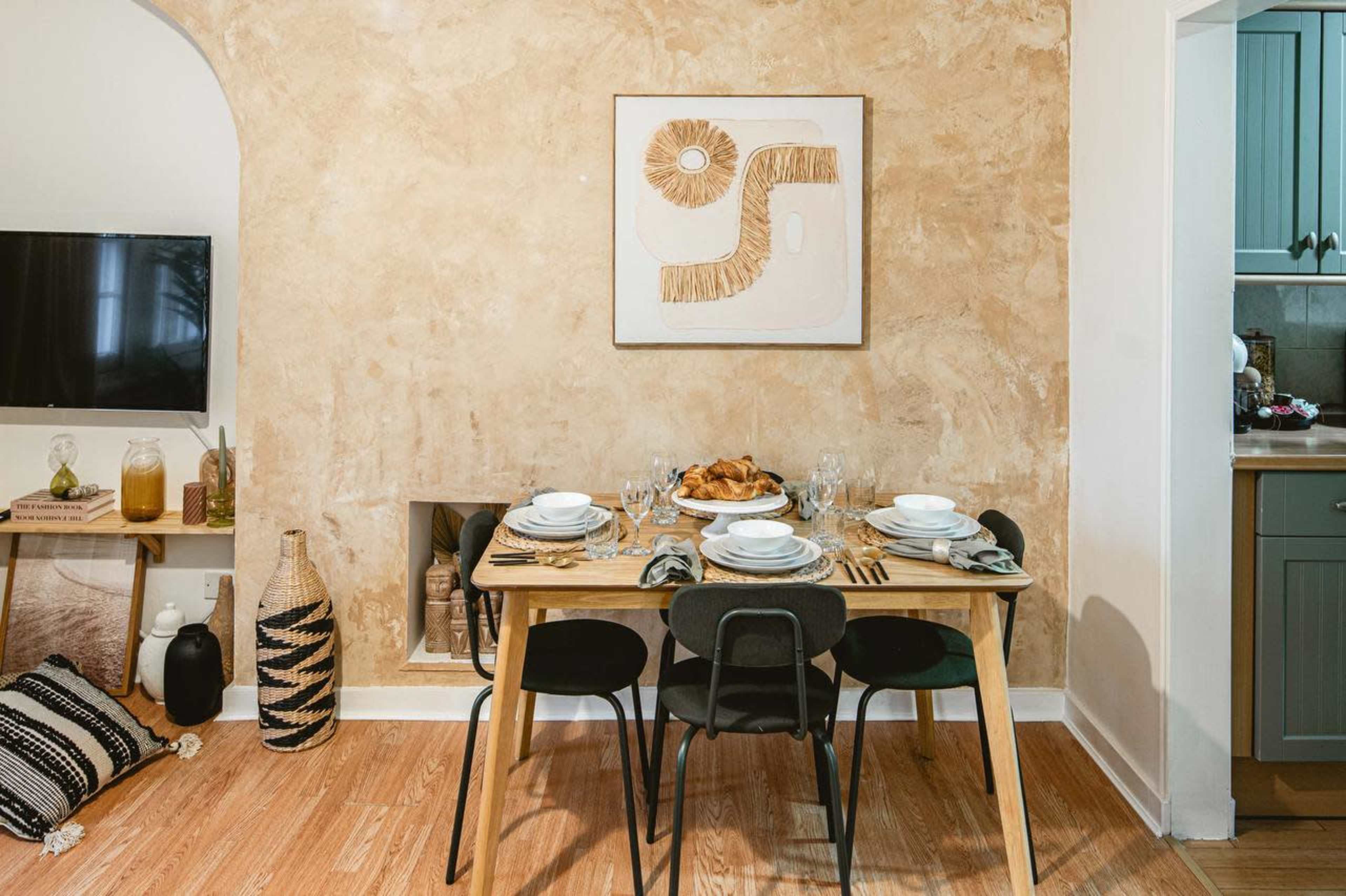 A wooden dining table is set with plates and glasses, positioned against a textured beige wall with abstract art, near a small kitchen area and a television.
