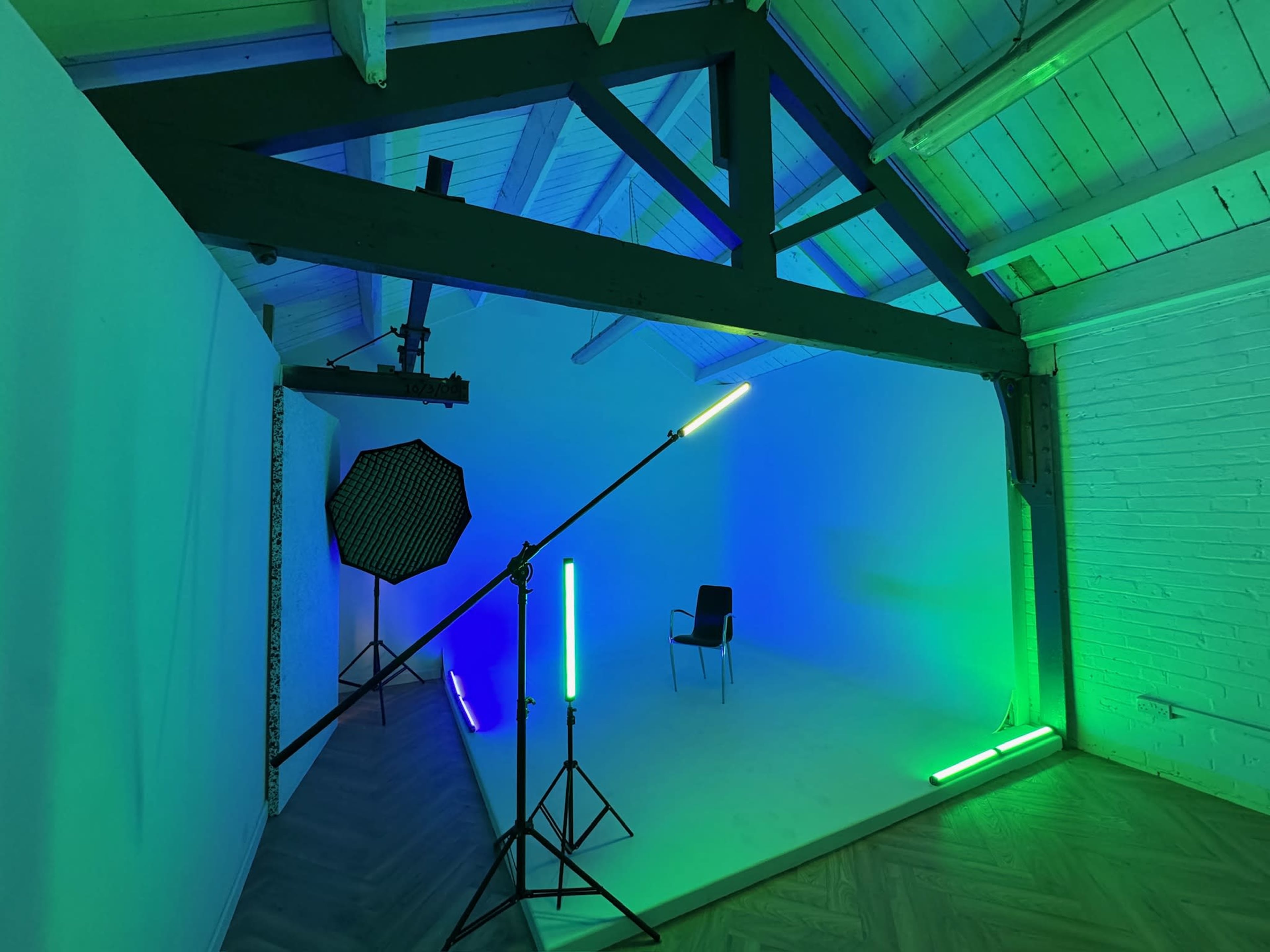 The image shows a photography studio with a white backdrop, a black chair, and various lighting equipment illuminating the space in green and blue tones.