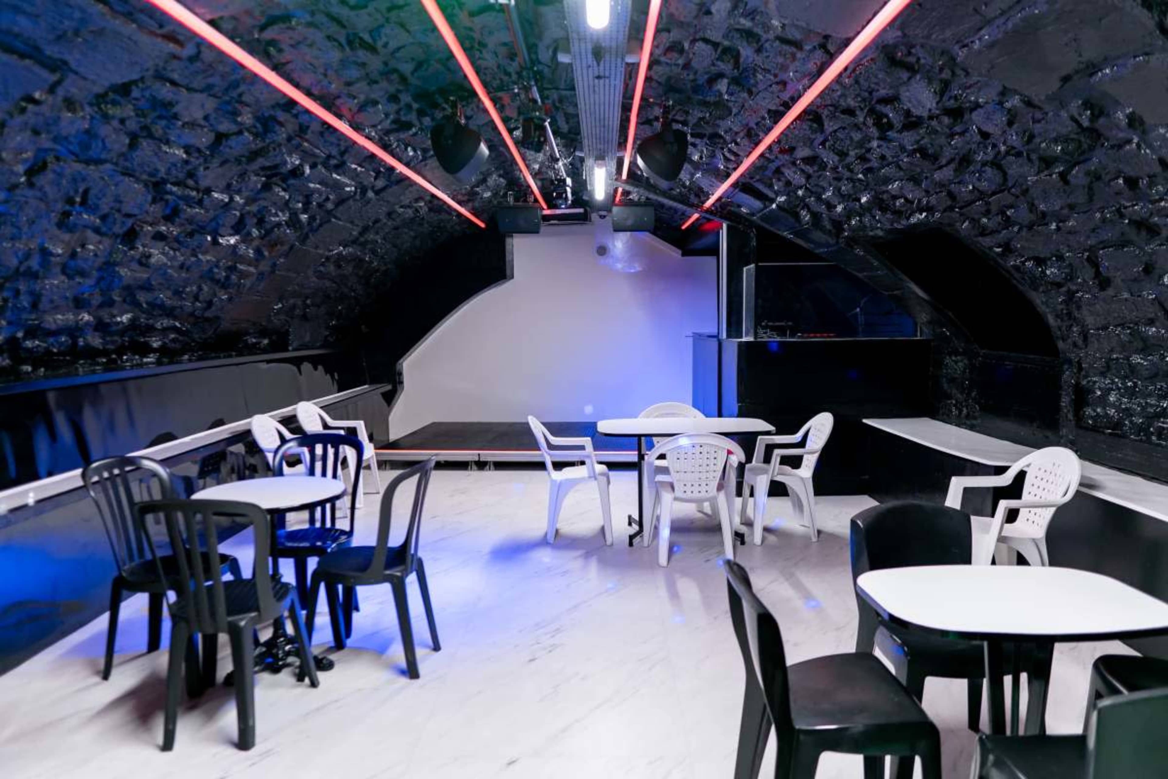 The image shows a modern lounge area with several white and black chairs and tables under a curved, textured ceiling featuring colorful neon lights.