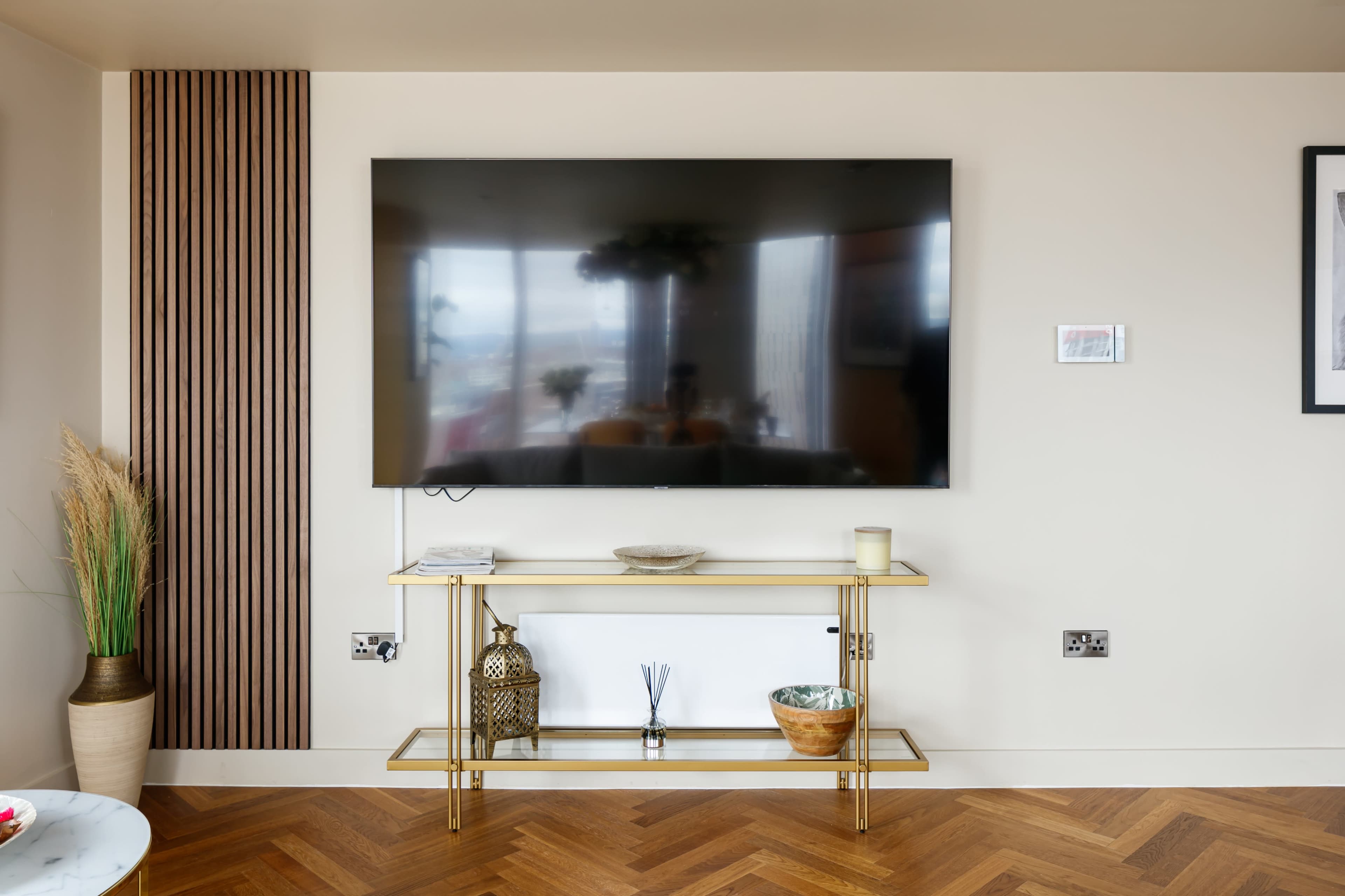 A large flat-screen television is mounted on a light-colored wall above a gold metal console table with decorative items, set against a backdrop of herringbone-patterned wood flooring.