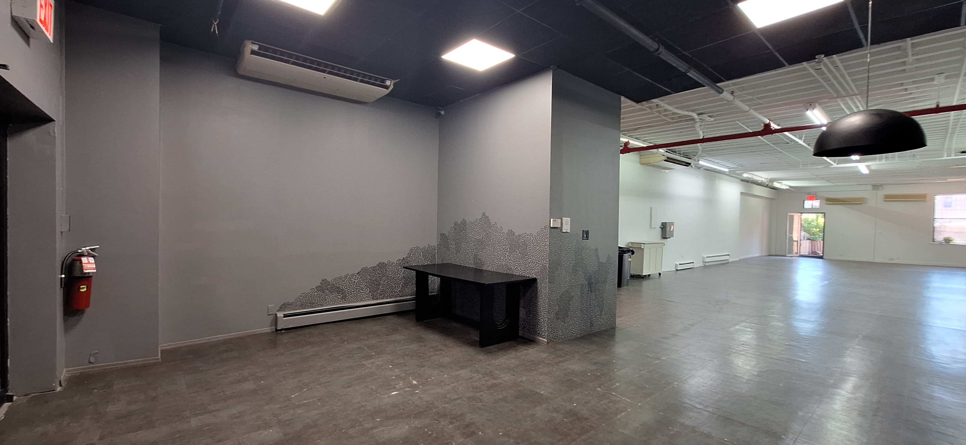 The image shows an empty interior space featuring a gray wall with a decorative pattern, a black table, and fluorescent ceiling lights.