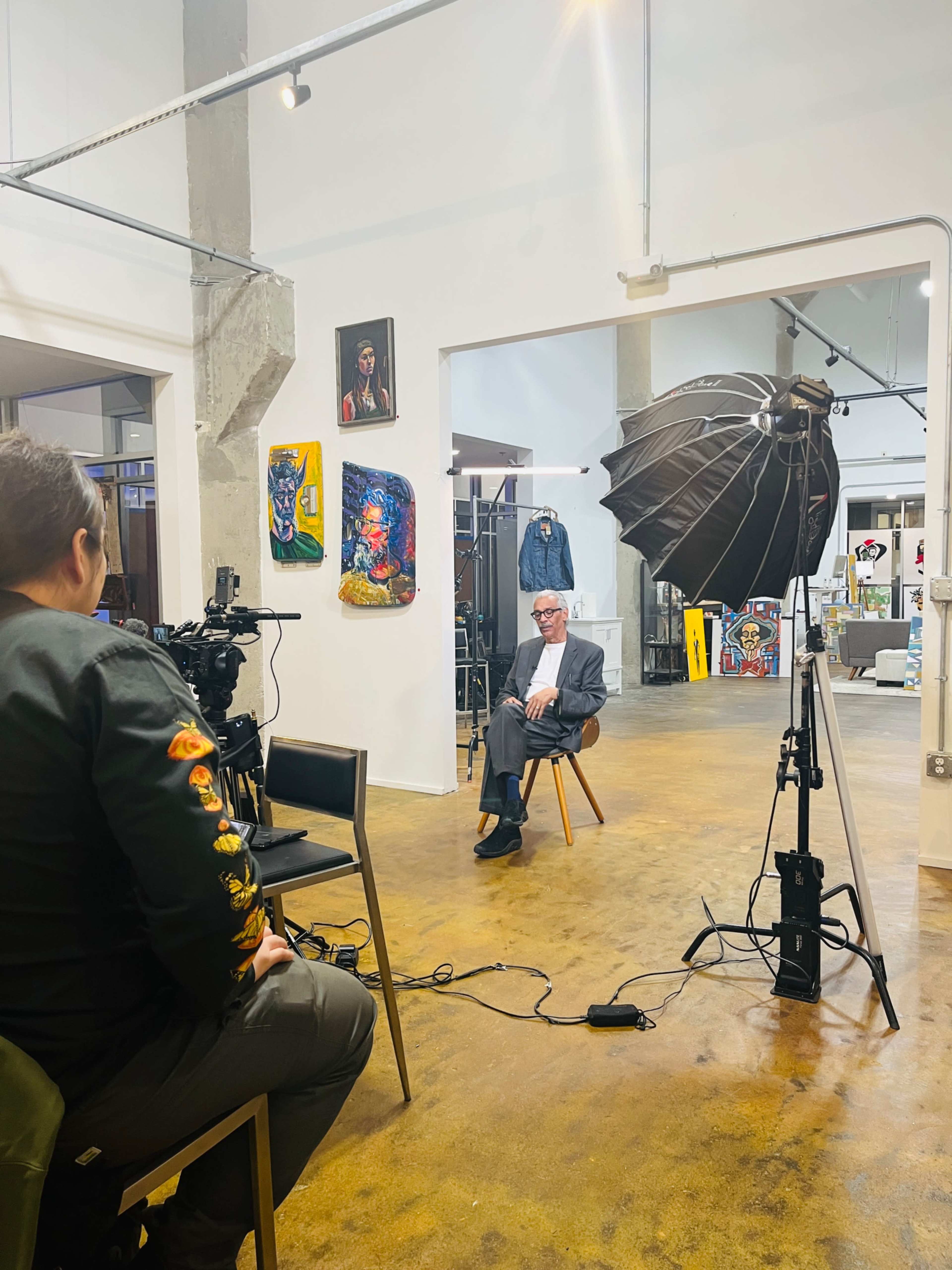 A man in a suit sits on a chair in an art gallery while a photographer prepares to take his picture with a large softbox light.