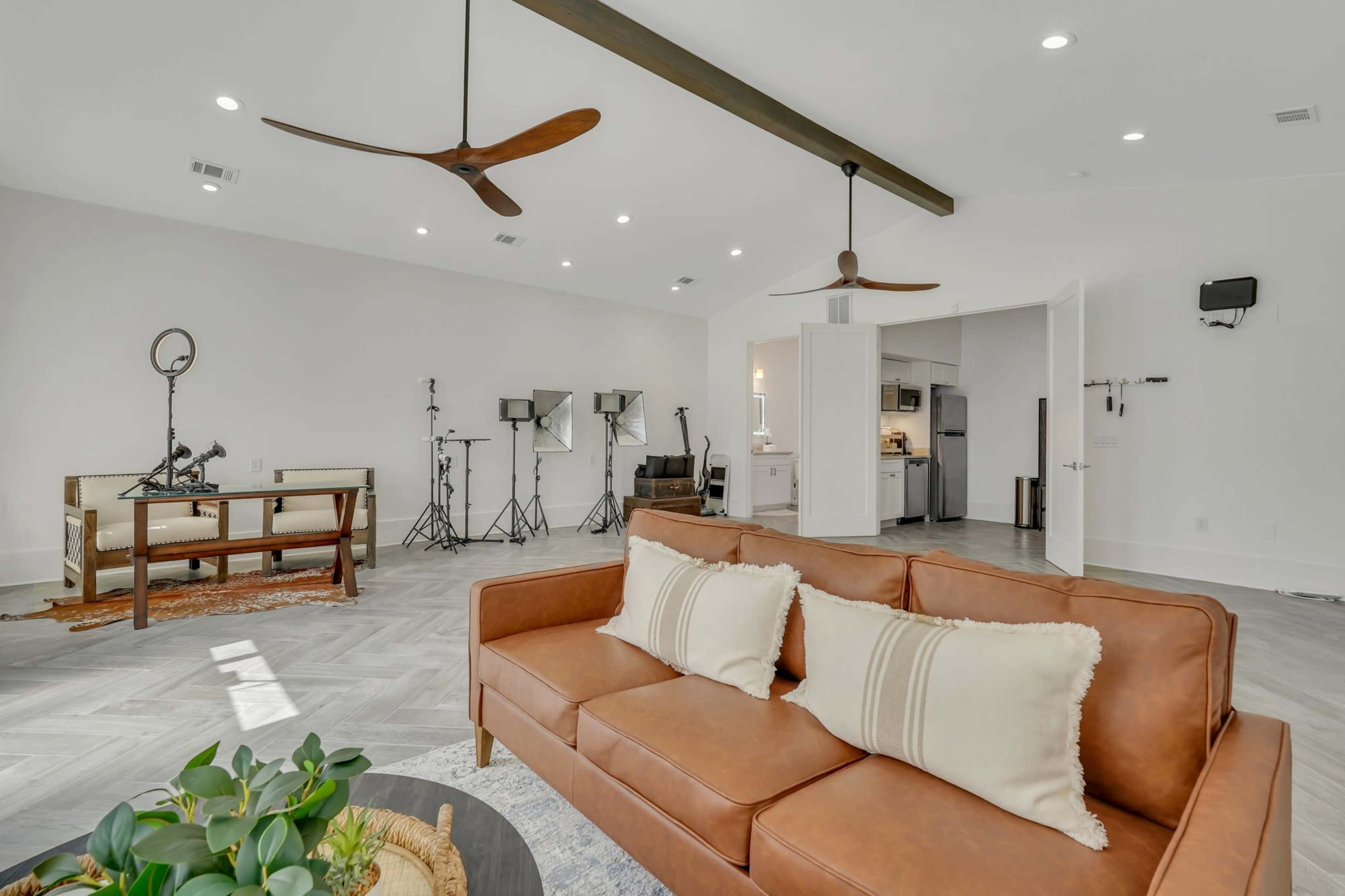 A modern living space with a brown leather sofa, a dining area, and a kitchen partially visible in the background, along with studio lighting equipment set up in the corner.