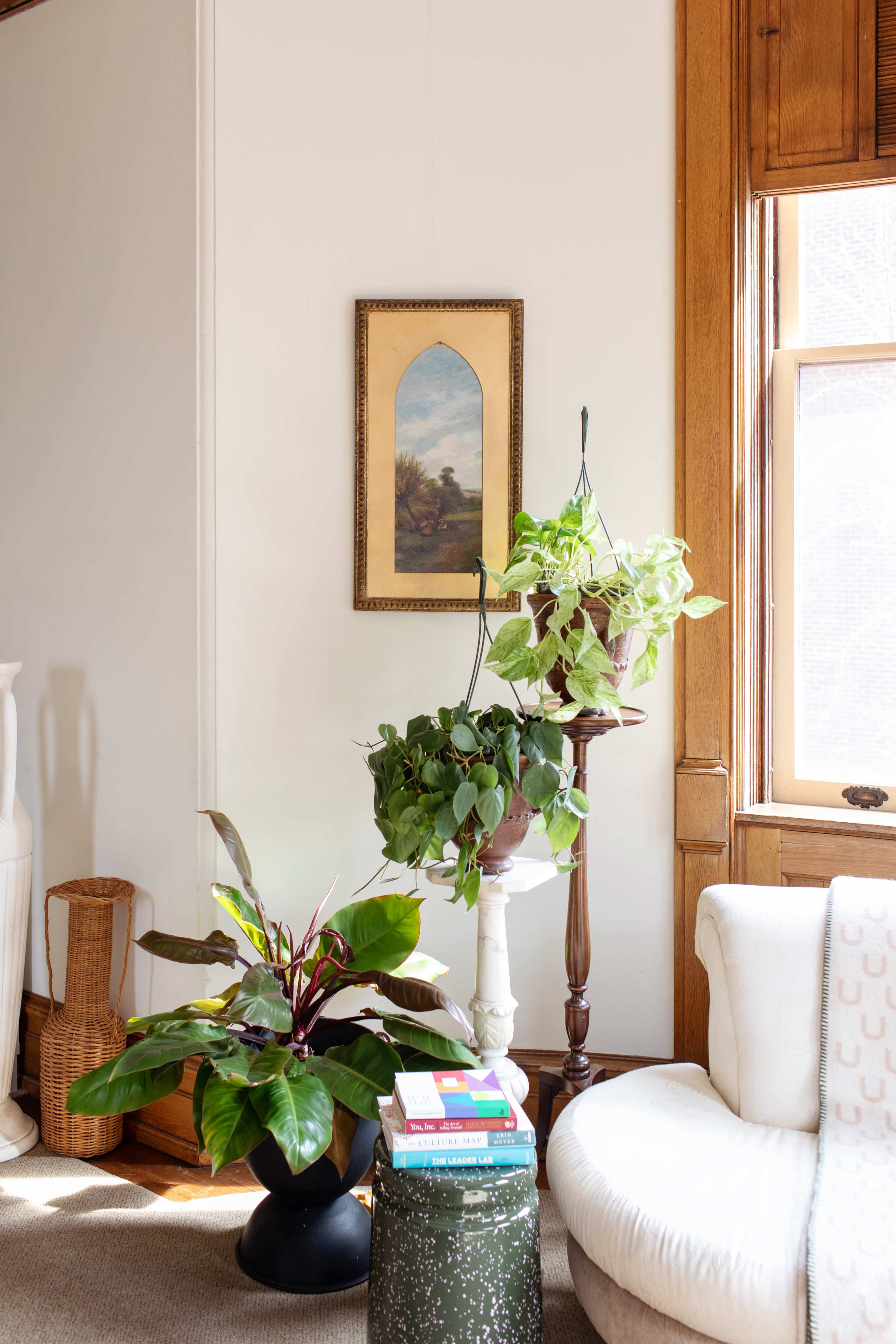 A bright living space featuring a potted plant on a pedestal, another plant in a hanging pot, a white couch, and a stack of books on a small table beside a large window.