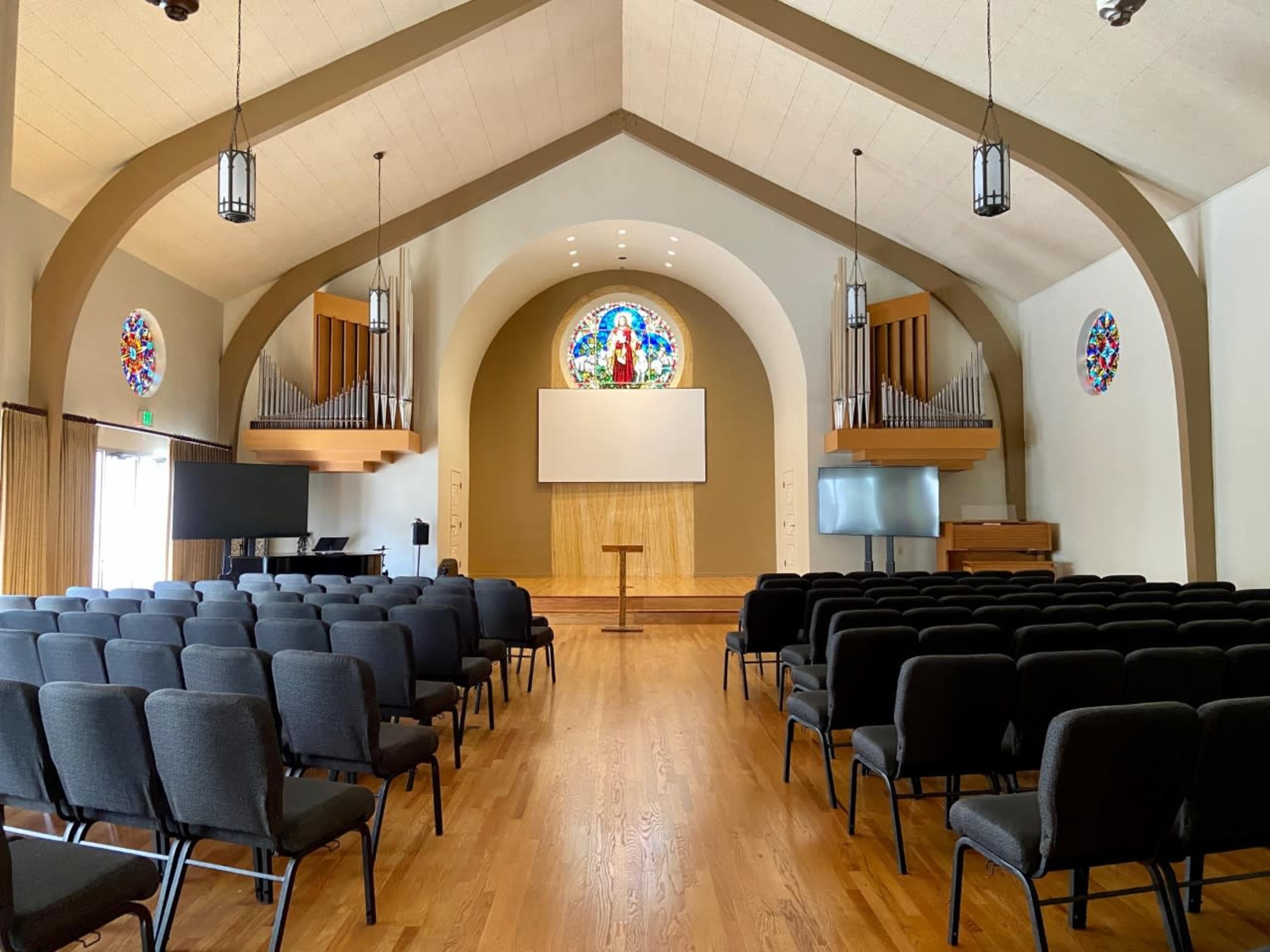 A spacious church interior with rows of black chairs facing a wooden podium and a large screen, flanked by stained glass windows and organ pipes.