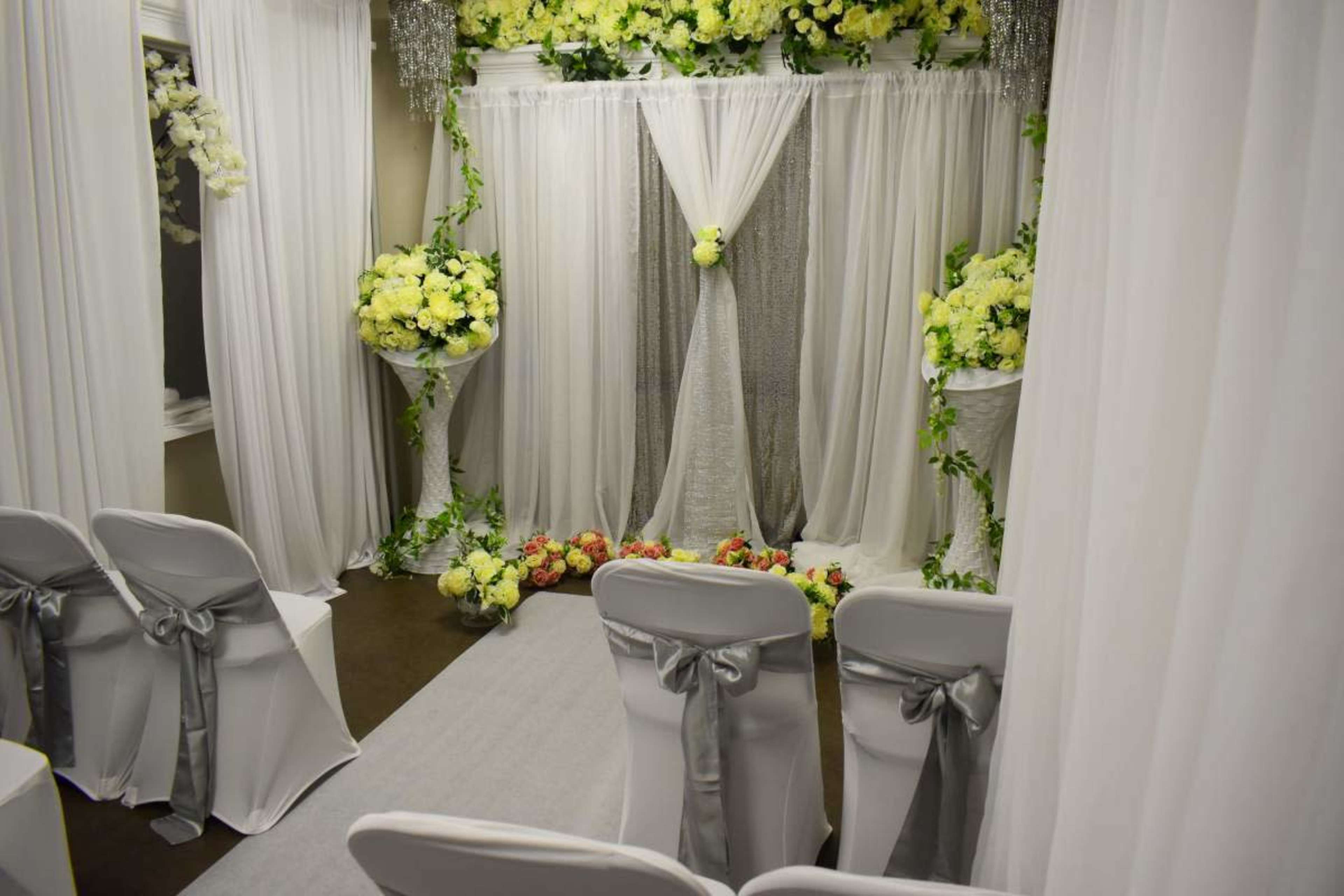 A decorated wedding ceremony space features white drapery, floral arrangements, and chairs arranged on a grey runner.