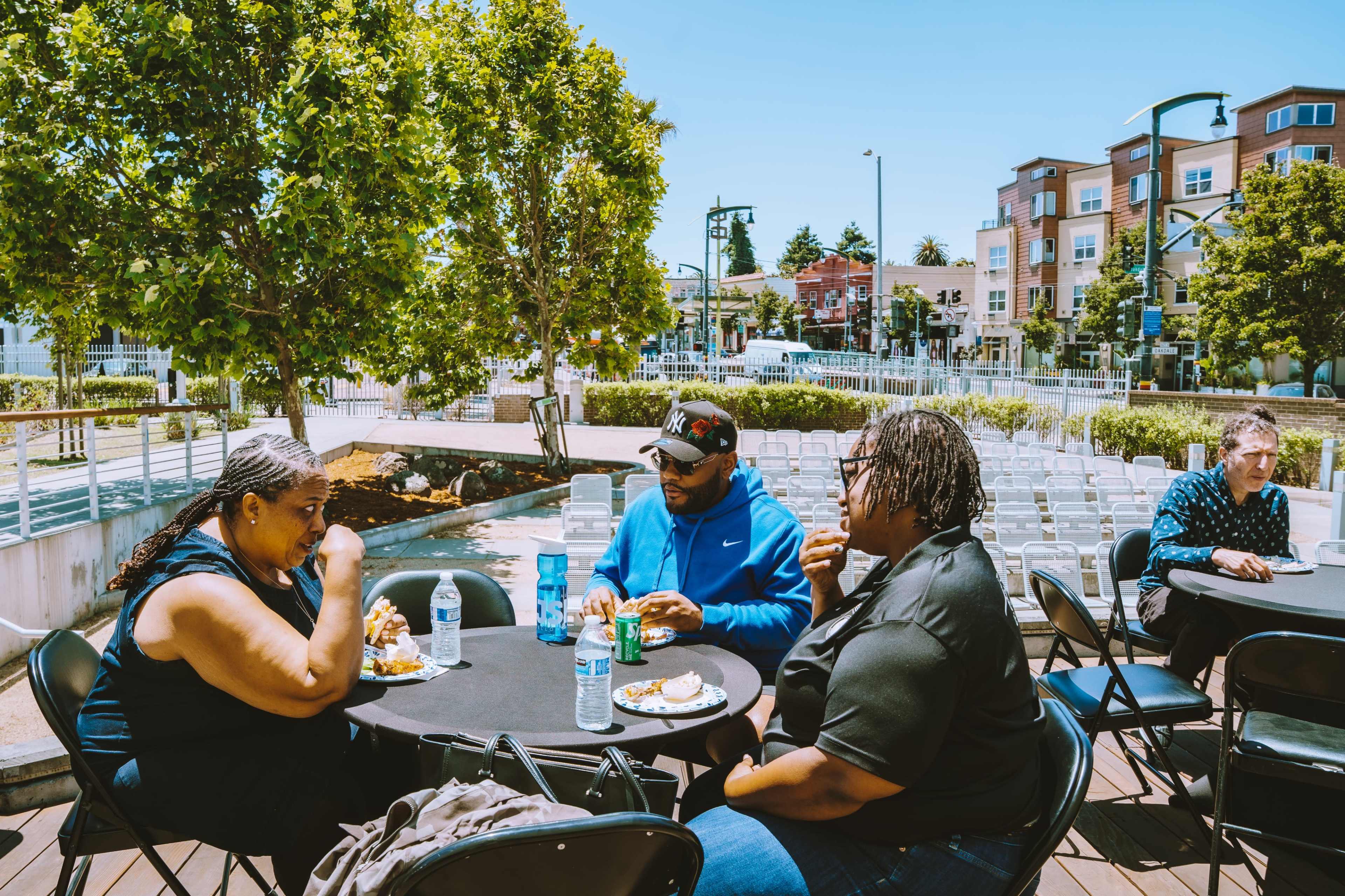 A group of four people is seated at a table outdoors, enjoying a meal with a view of a nearby water feature and buildings in the background.