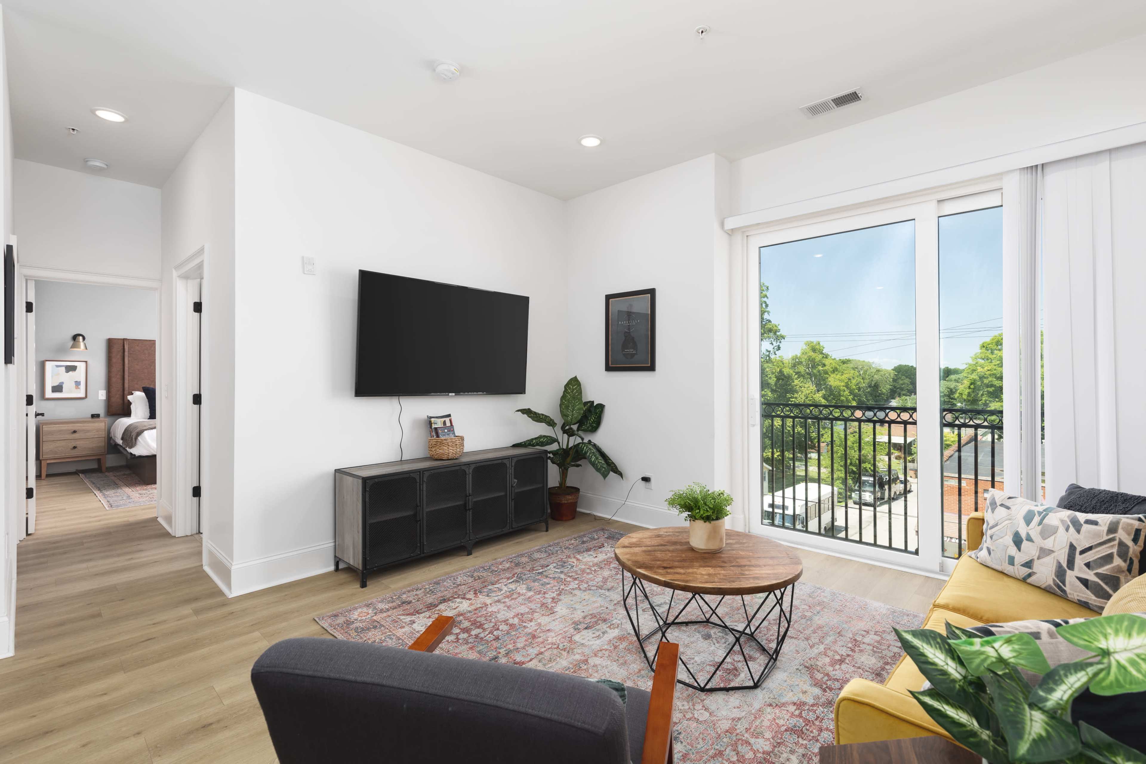 A modern living room features a large TV mounted on the wall, a cozy yellow sofa, and a round coffee table on a patterned rug, with a view of a balcony through sliding glass doors.