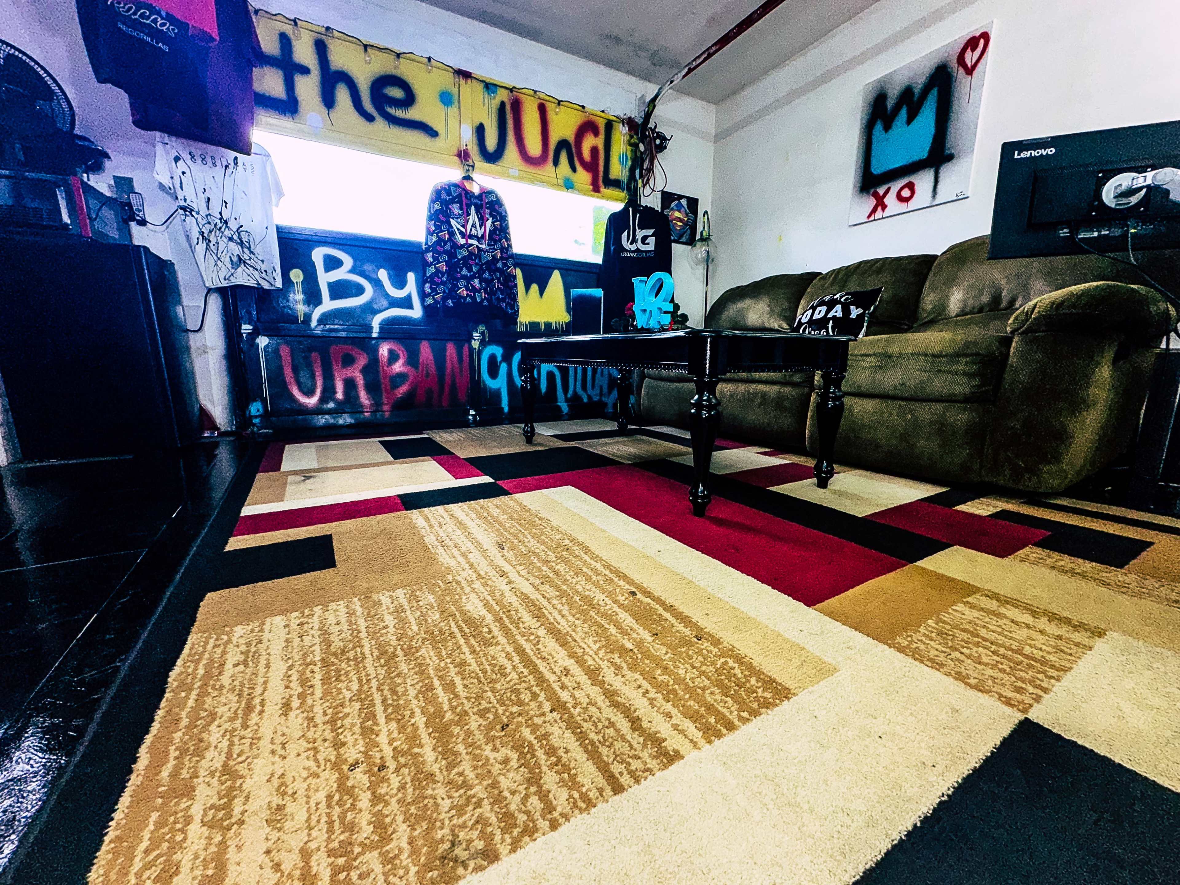 A room with a patterned rug, a green couch, colorful graffiti on the walls, and clothes hanging on a rack.
