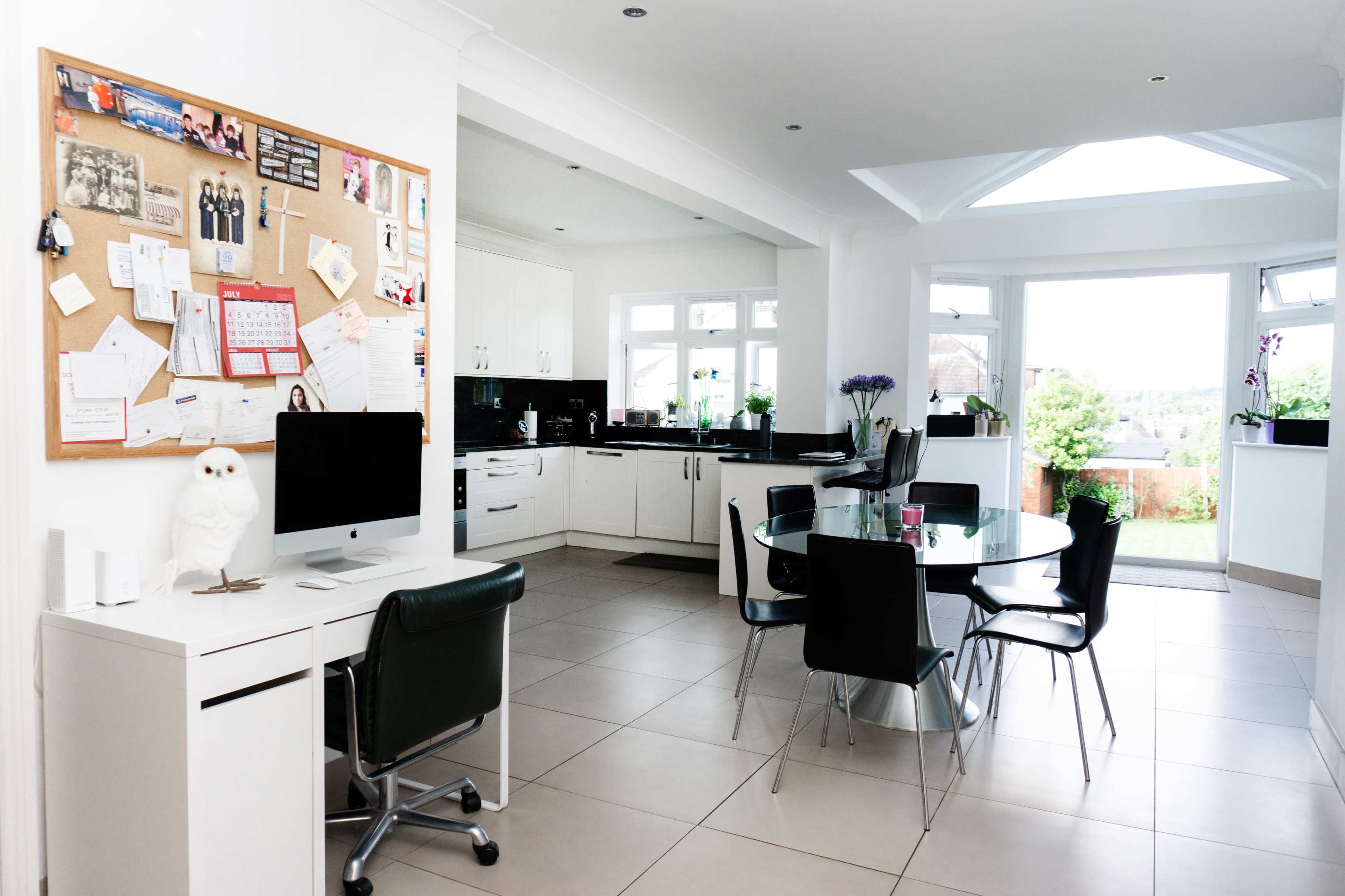 A modern kitchen and dining area features a bulletin board, a computer setup, a circular dining table, and large windows that provide natural light.