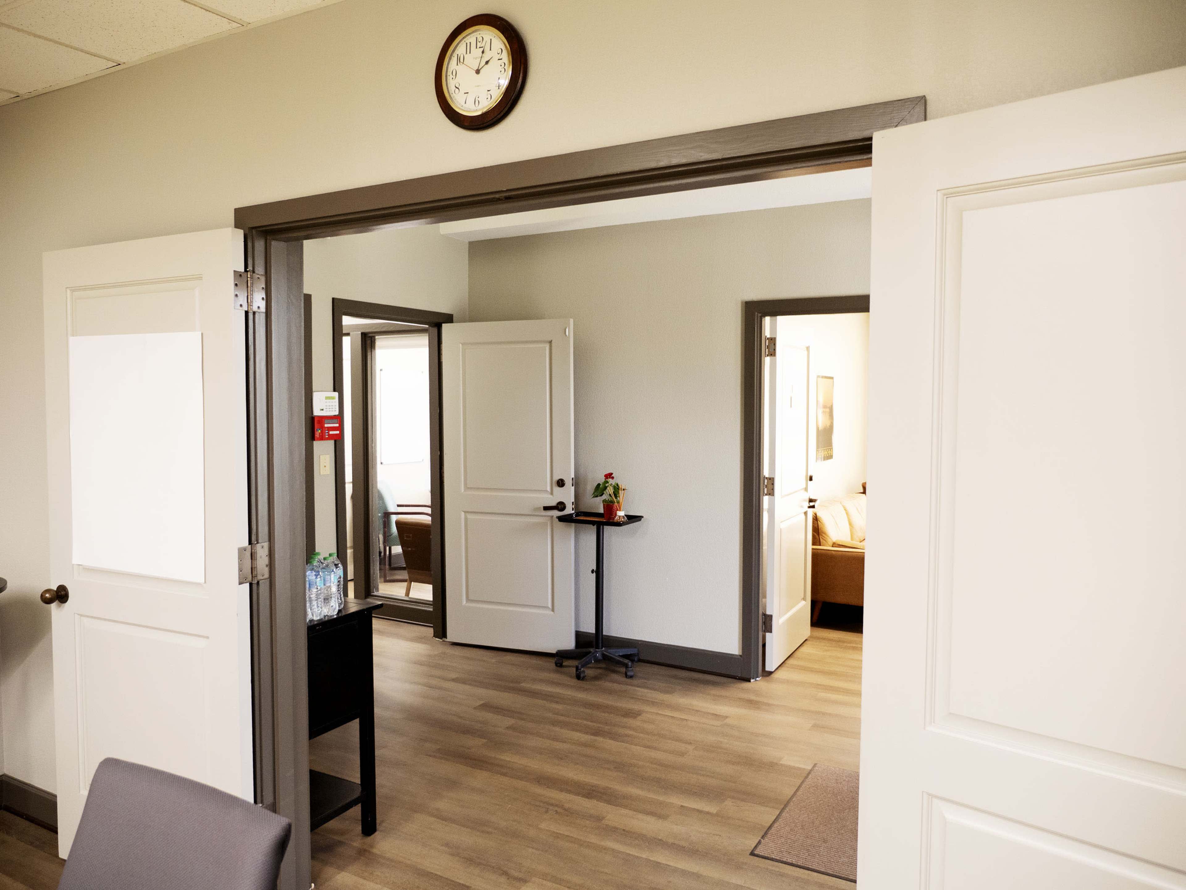 The image shows an interior space featuring an open doorway with a clock on the wall, leading to two separate rooms with simple furnishings.