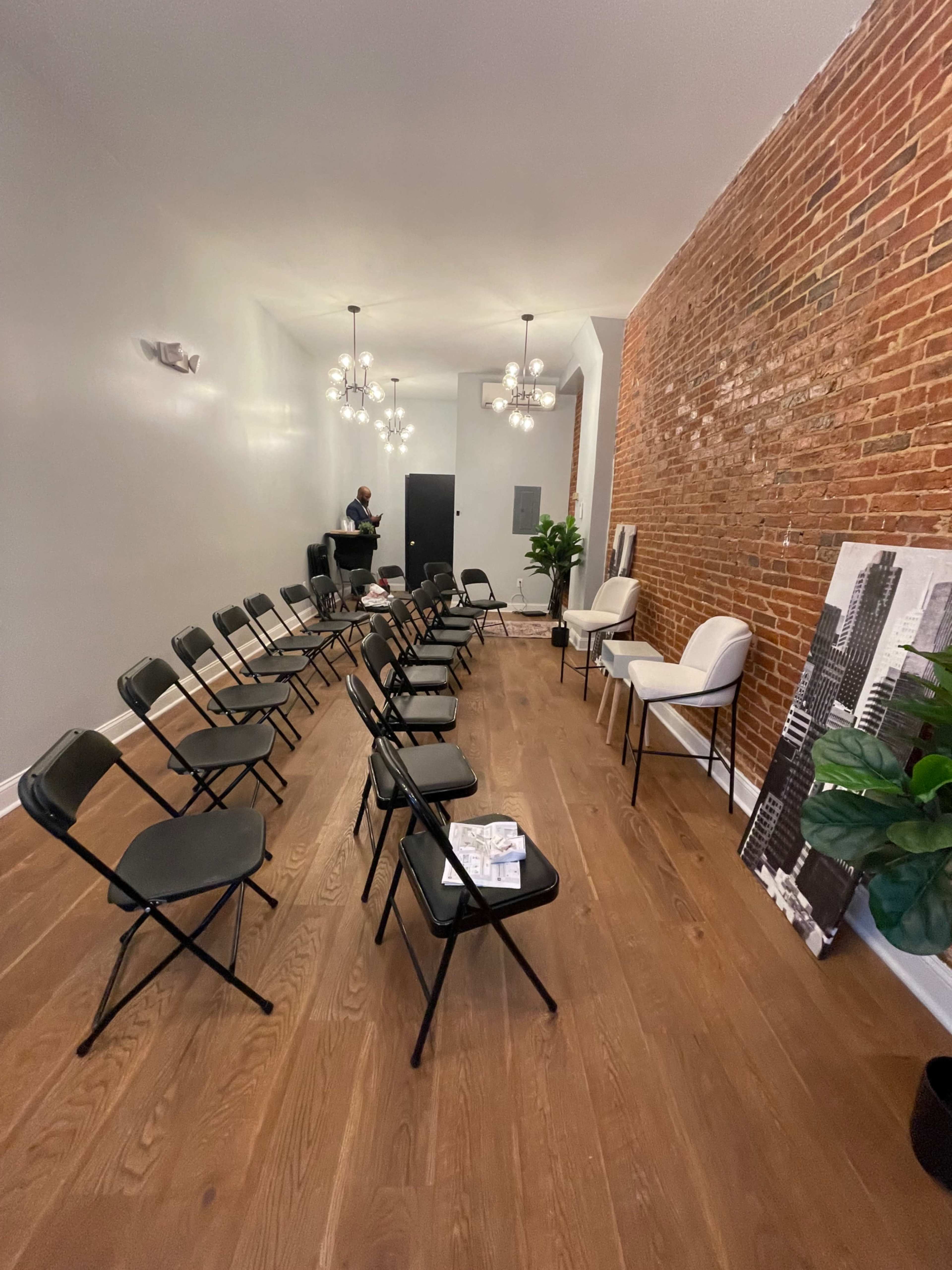 A waiting area with two rows of black folding chairs facing a wall adorned with exposed brick and decorative lighting fixtures.