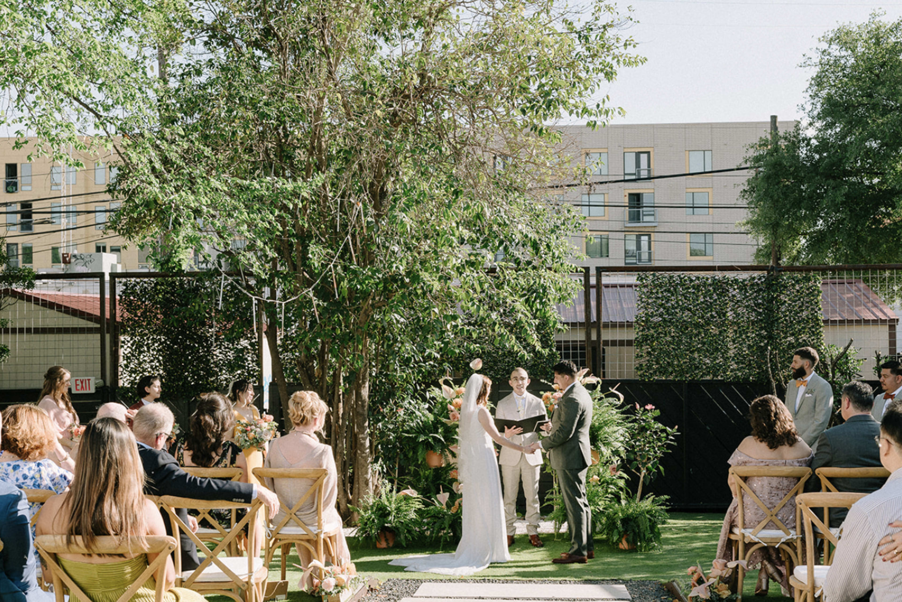 A couple exchanges vows during a wedding ceremony in a garden setting surrounded by guests seated on wooden chairs.