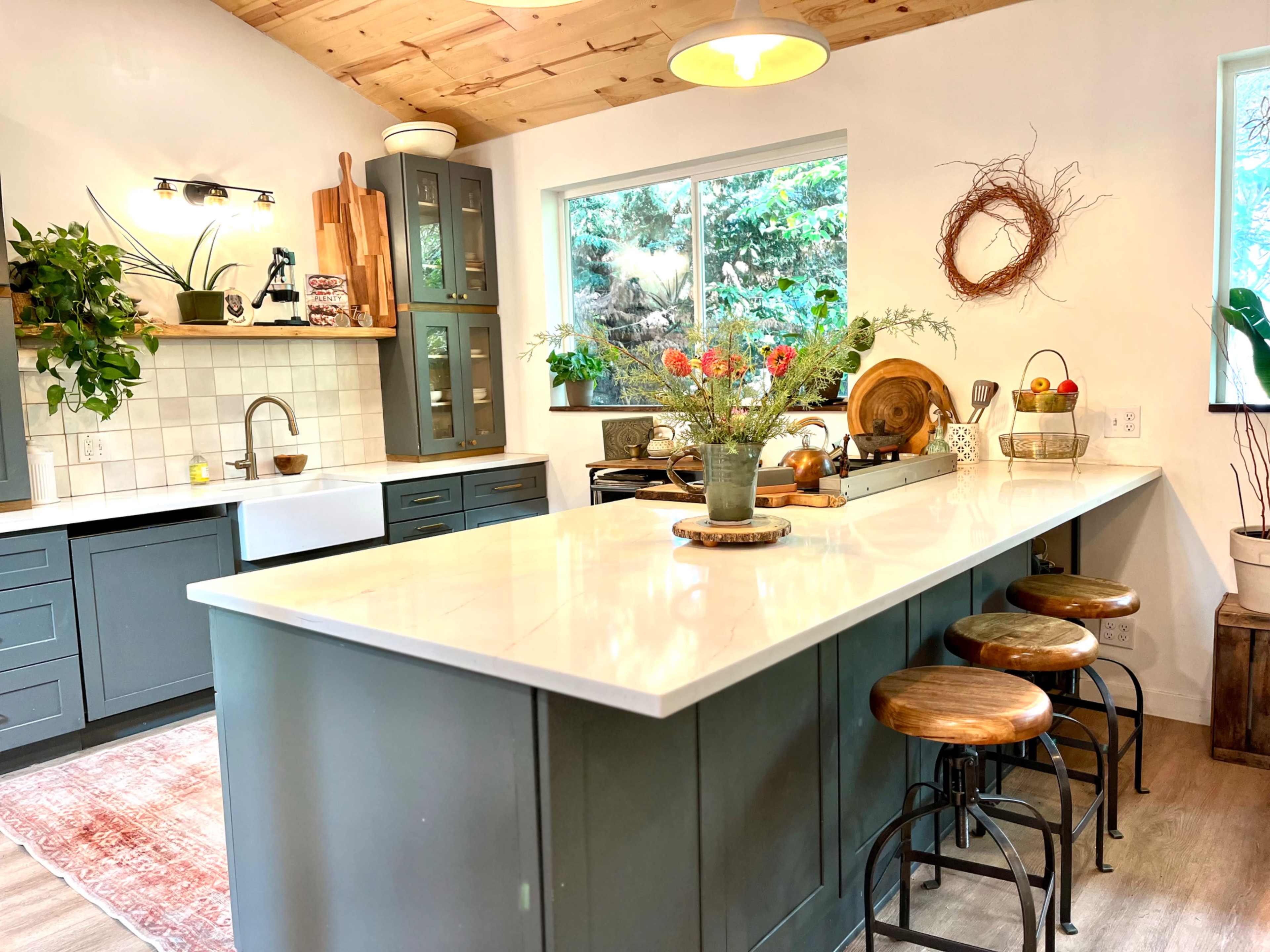 The image shows a modern kitchen featuring a central island with wooden stools, green cabinets, a farmhouse sink, and large windows allowing natural light to fill the space.