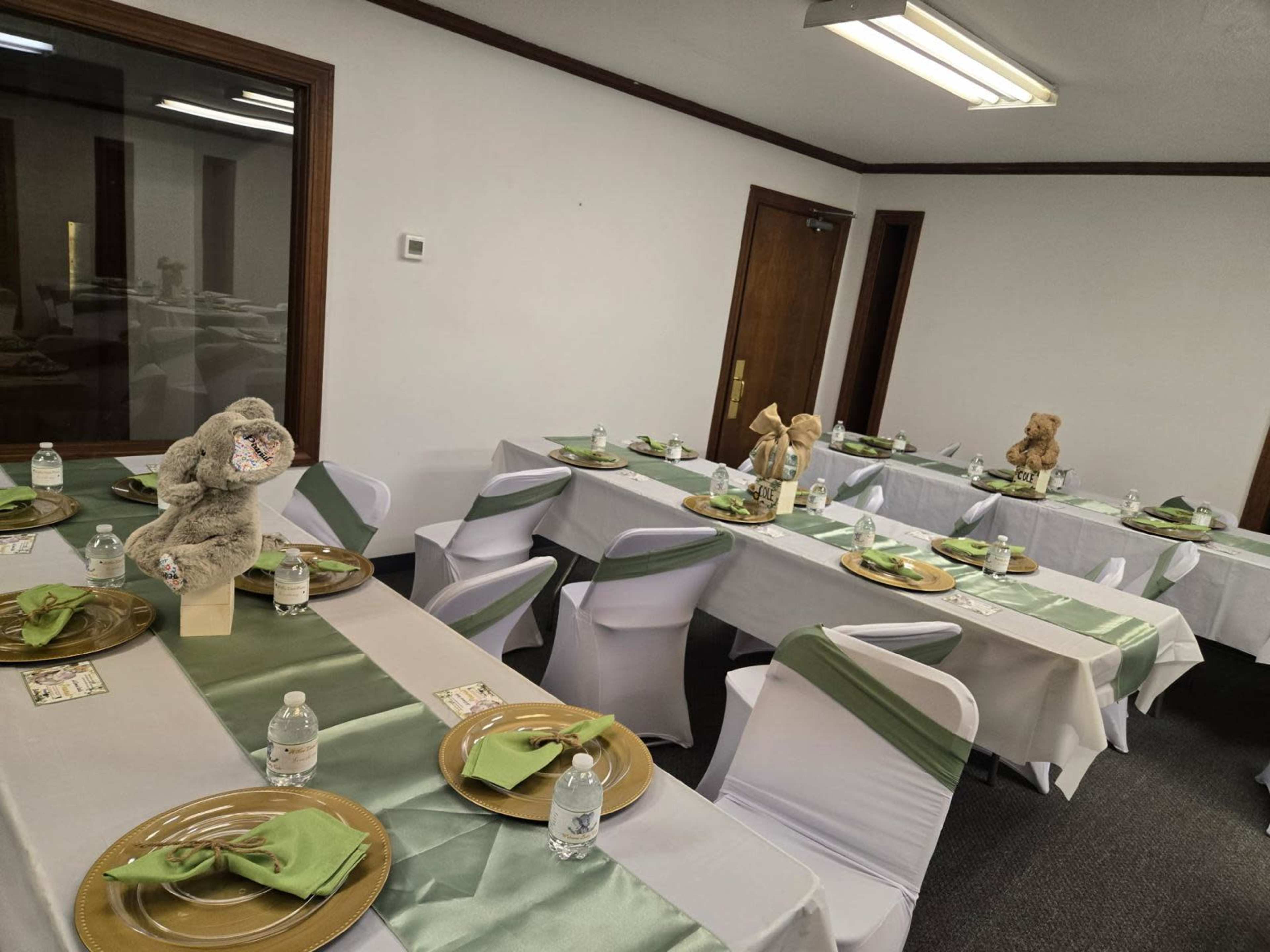 The image shows a spacious room set up with tables covered in white tablecloths, adorned with green sashes, and arranged with plates and bottled water, along with teddy bear centerpieces.