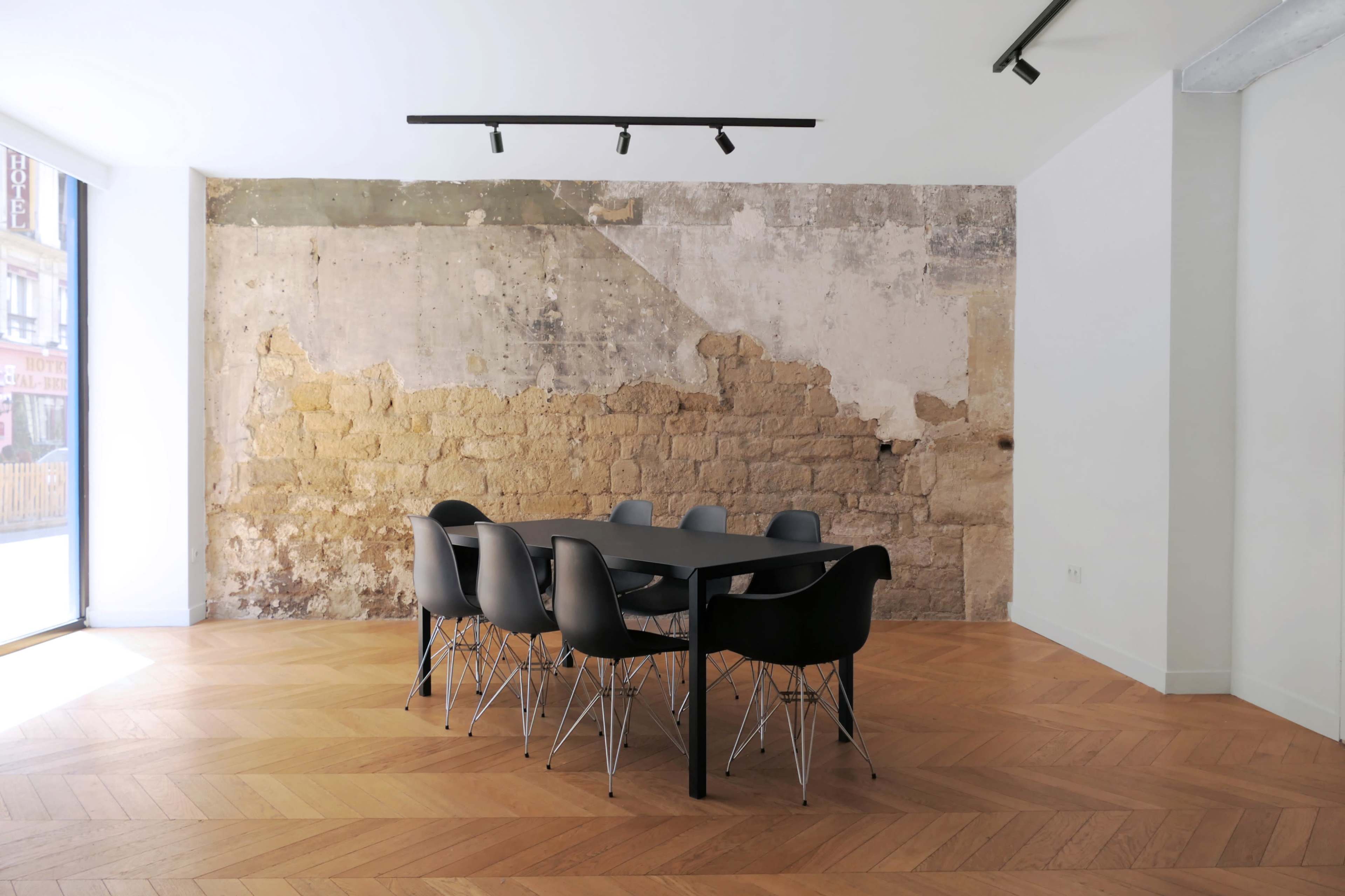 A modern dining area features a large black table surrounded by sleek black chairs, set against a rustic textured wall and wooden herringbone flooring.