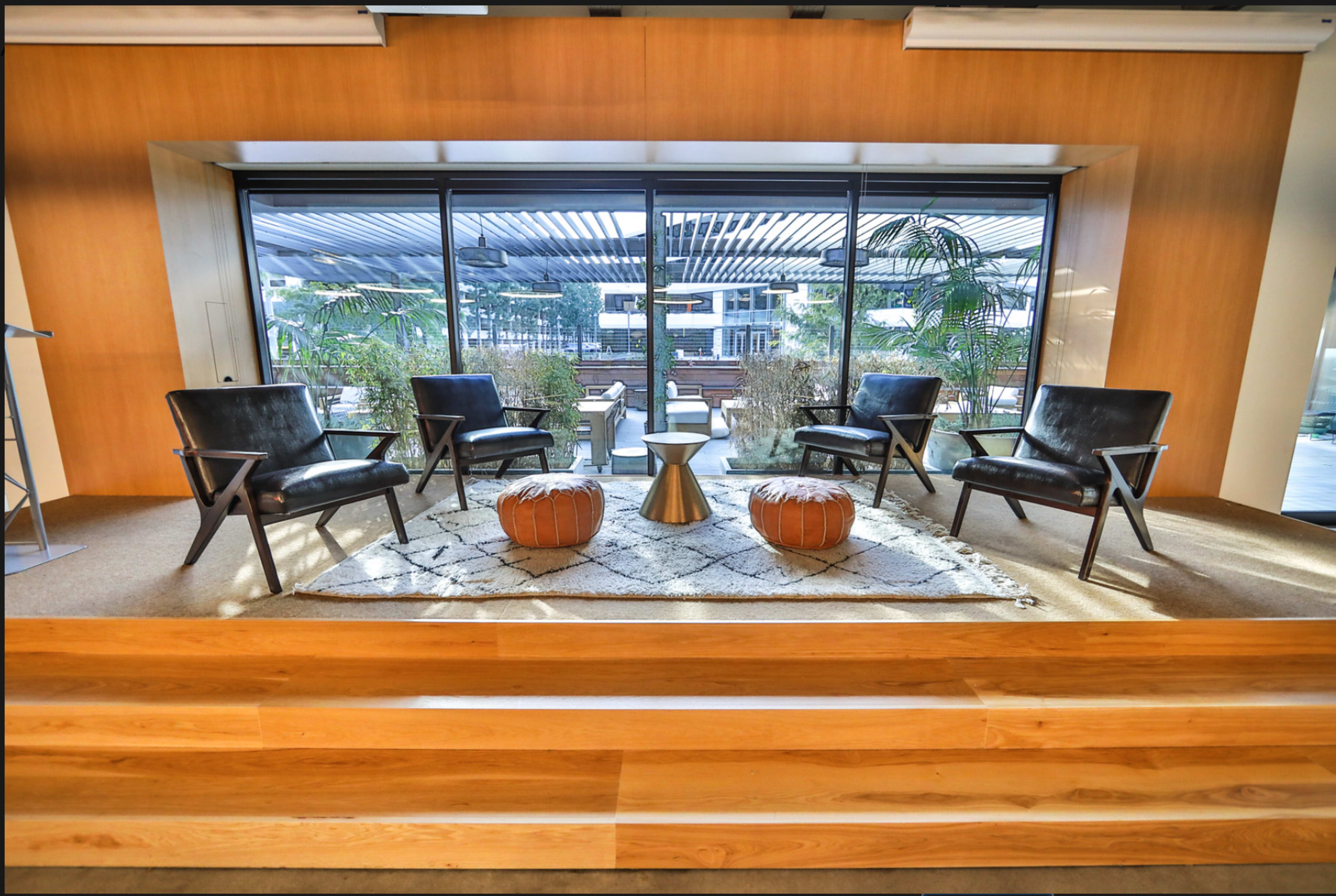 The image shows a modern, minimalist setting with four black chairs arranged around a central table on a raised platform, overlooking an outdoor area through large glass windows.
