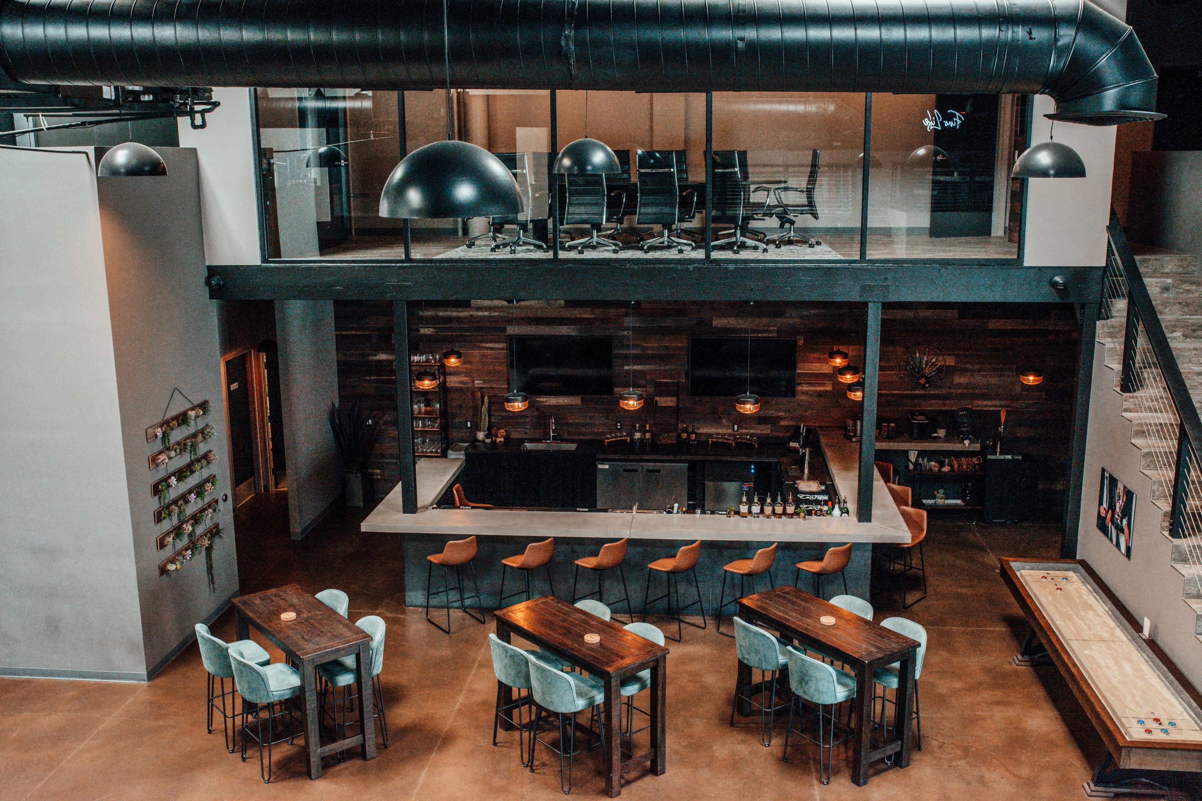 A modern bar area with wooden tables and chairs, a long counter with bar stools, and a lofted space above featuring office-style seating arrangements.