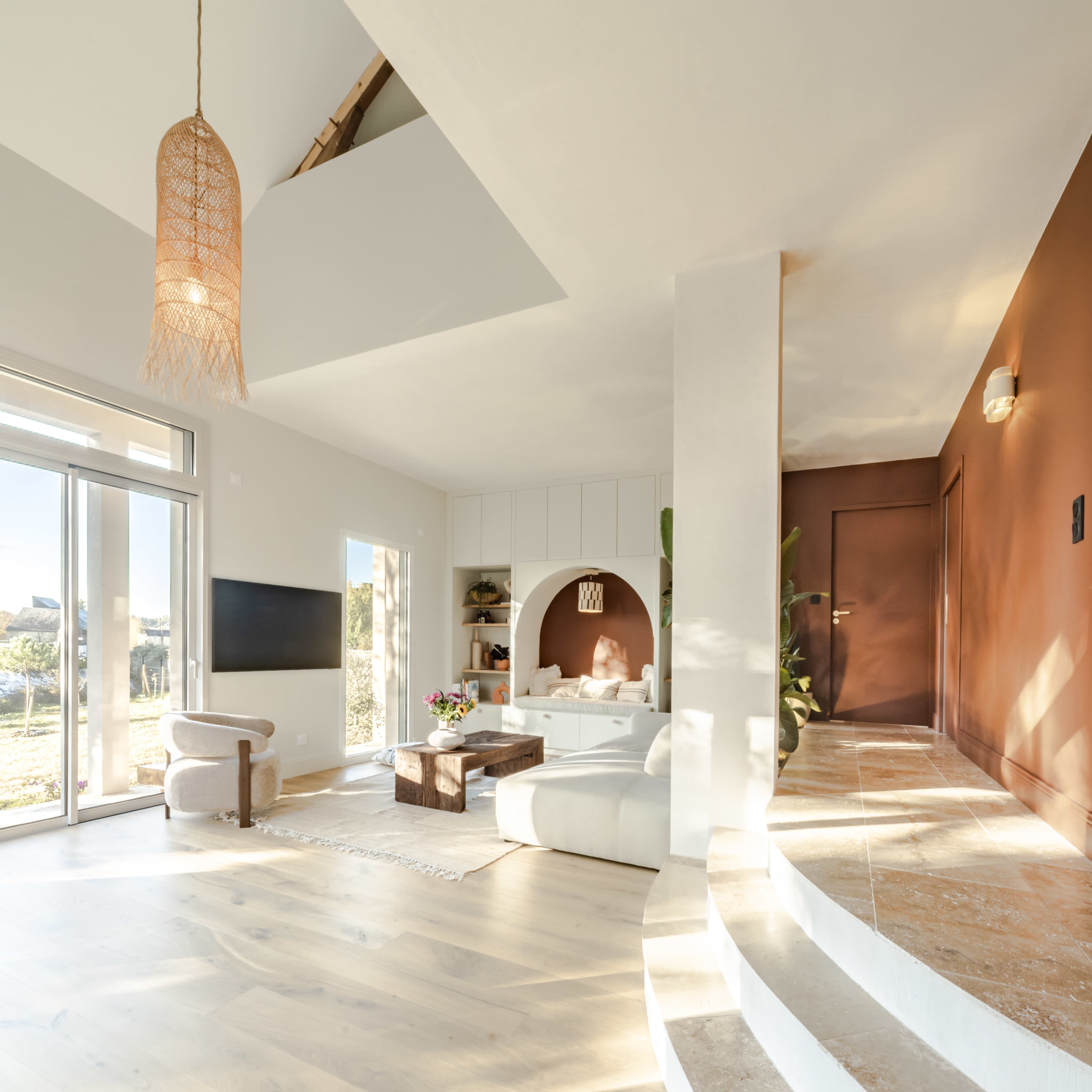 The image shows a modern living room with large windows, light wooden floors, and a circular seating area next to a wall with a geometric design.
