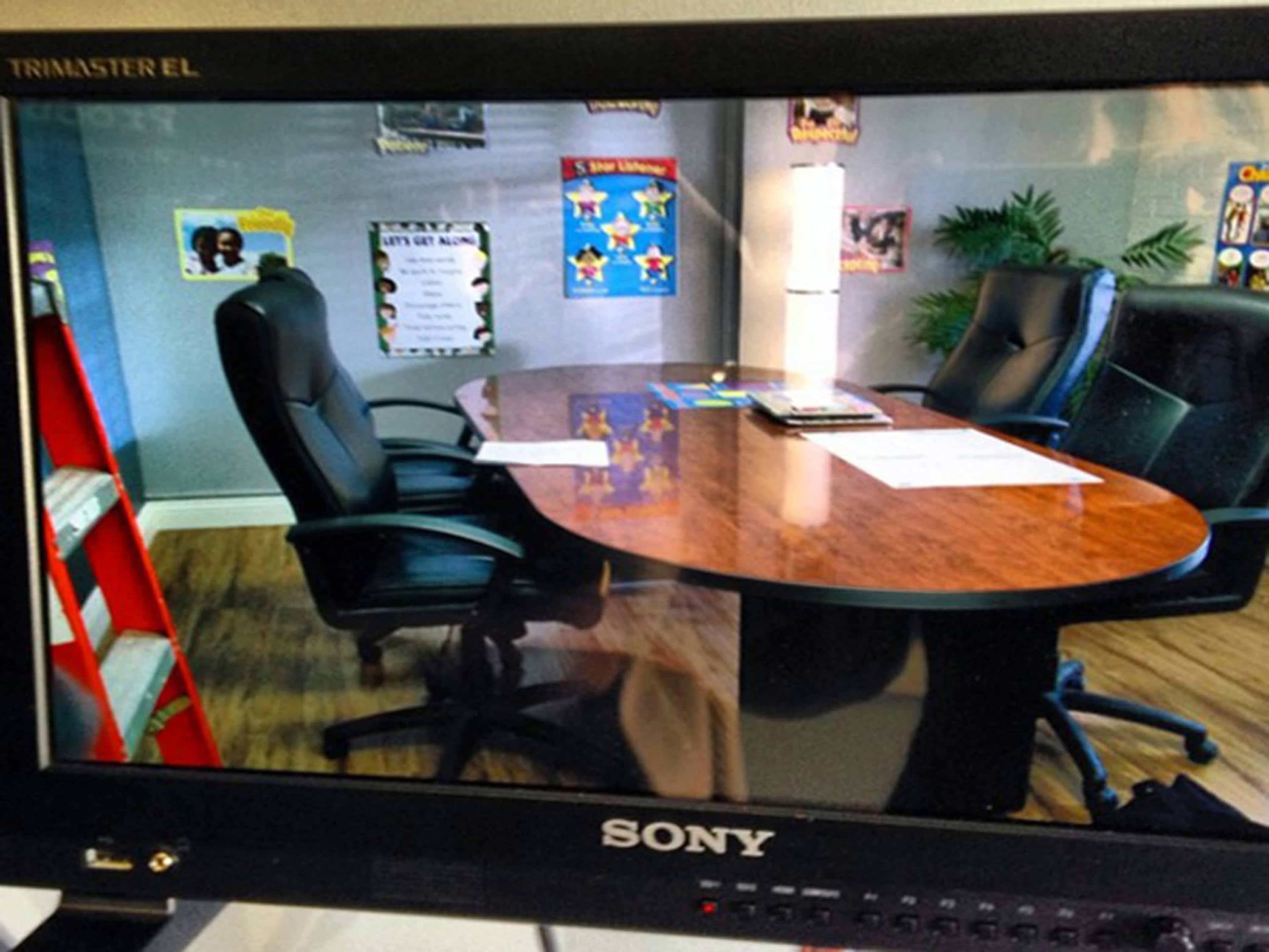 A conference room is displayed on a monitor, featuring an oval wooden table and two black chairs, surrounded by posters on the walls.