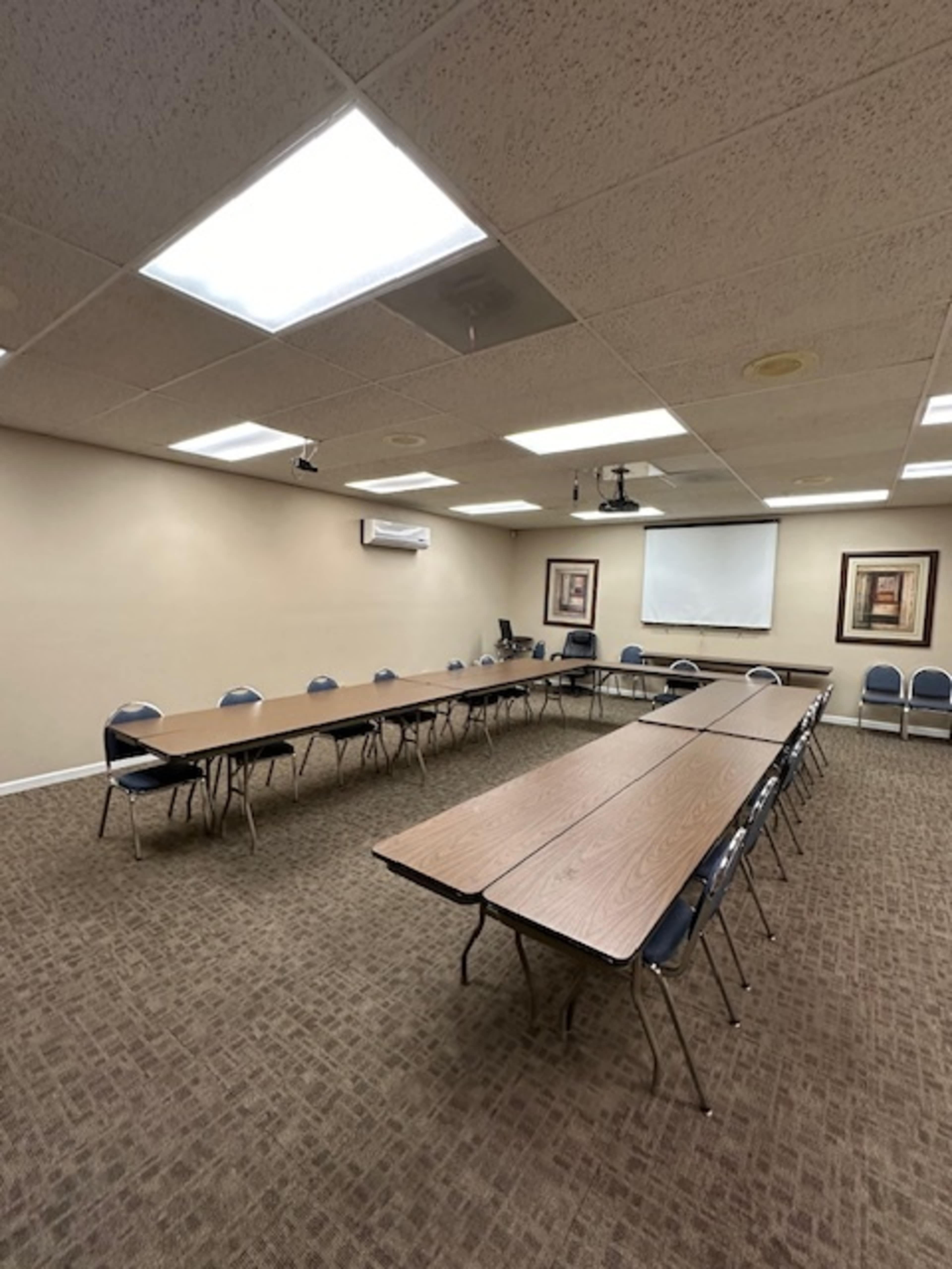 A conference room is set up with long tables arranged in a U-shape, surrounded by chairs, and features a screen on the front wall.