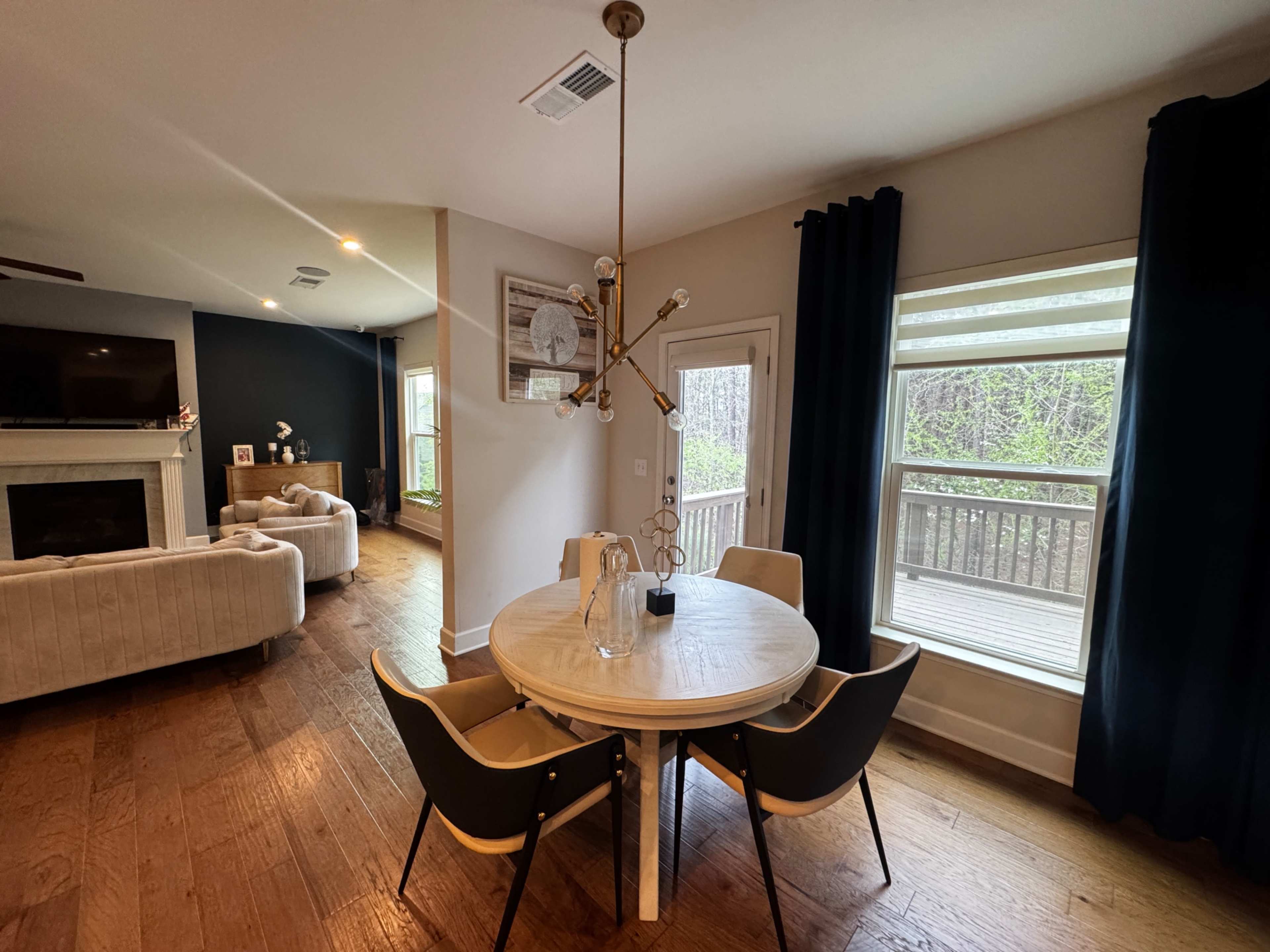 The image shows a dining area with a round wooden table and four chairs, illuminated by a modern light fixture, adjacent to a living space with a large couch and sliding glass doors leading to a deck.
