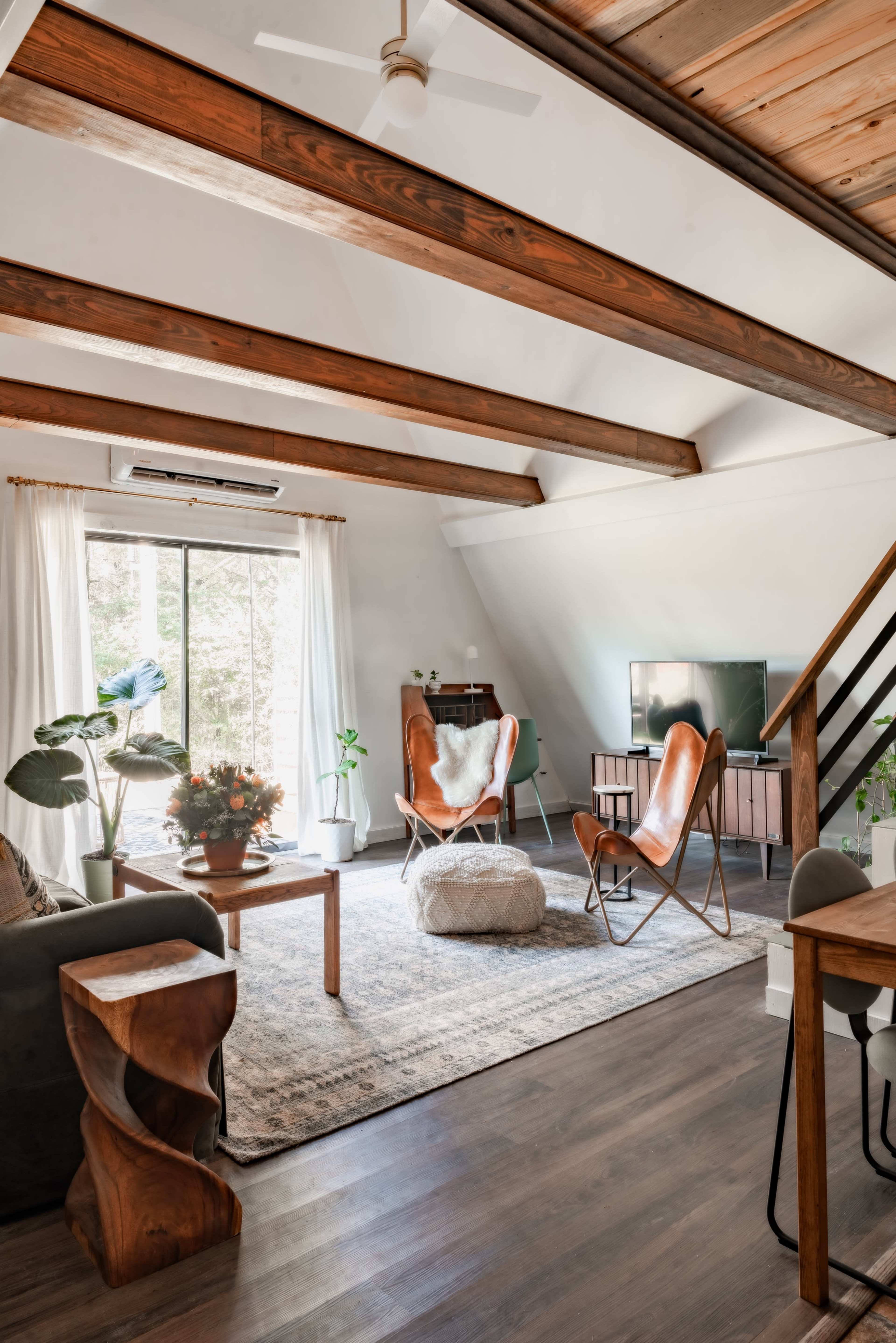 A cozy living room with wooden beams, a large window, a television, and a mix of modern and natural decor, including potted plants and stylish furniture.