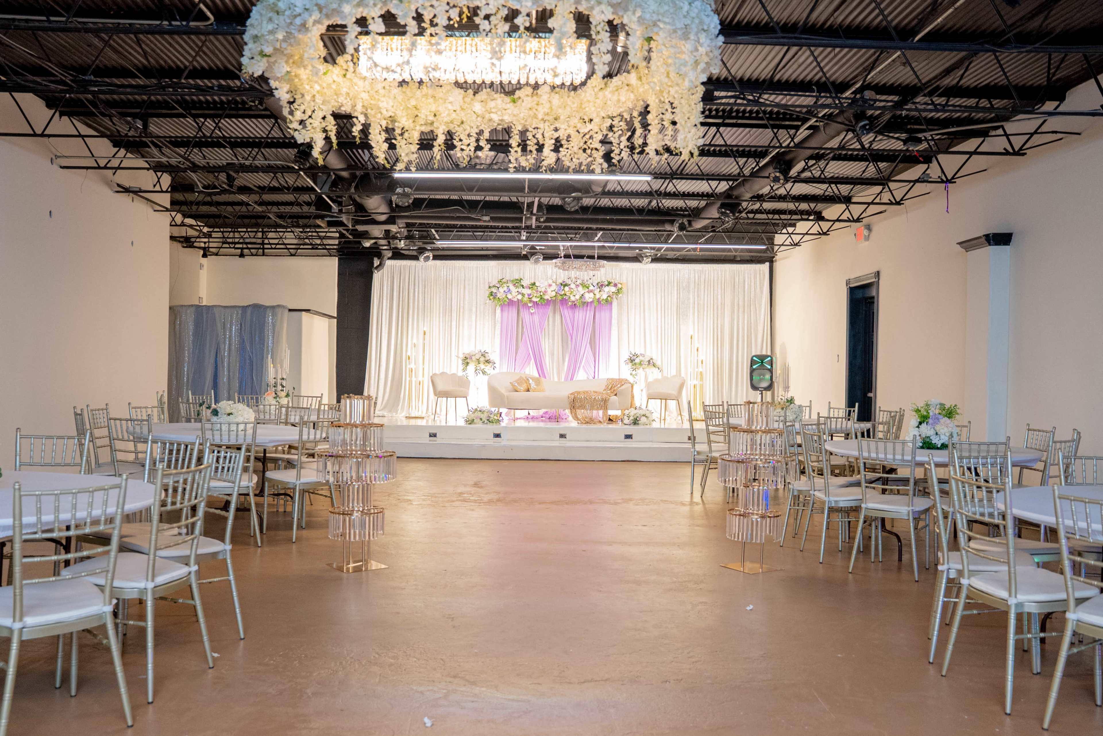 The image depicts a banquet hall set up for an event, featuring gold-accented chairs and round tables arranged around a stage adorned with floral decorations and drapery.