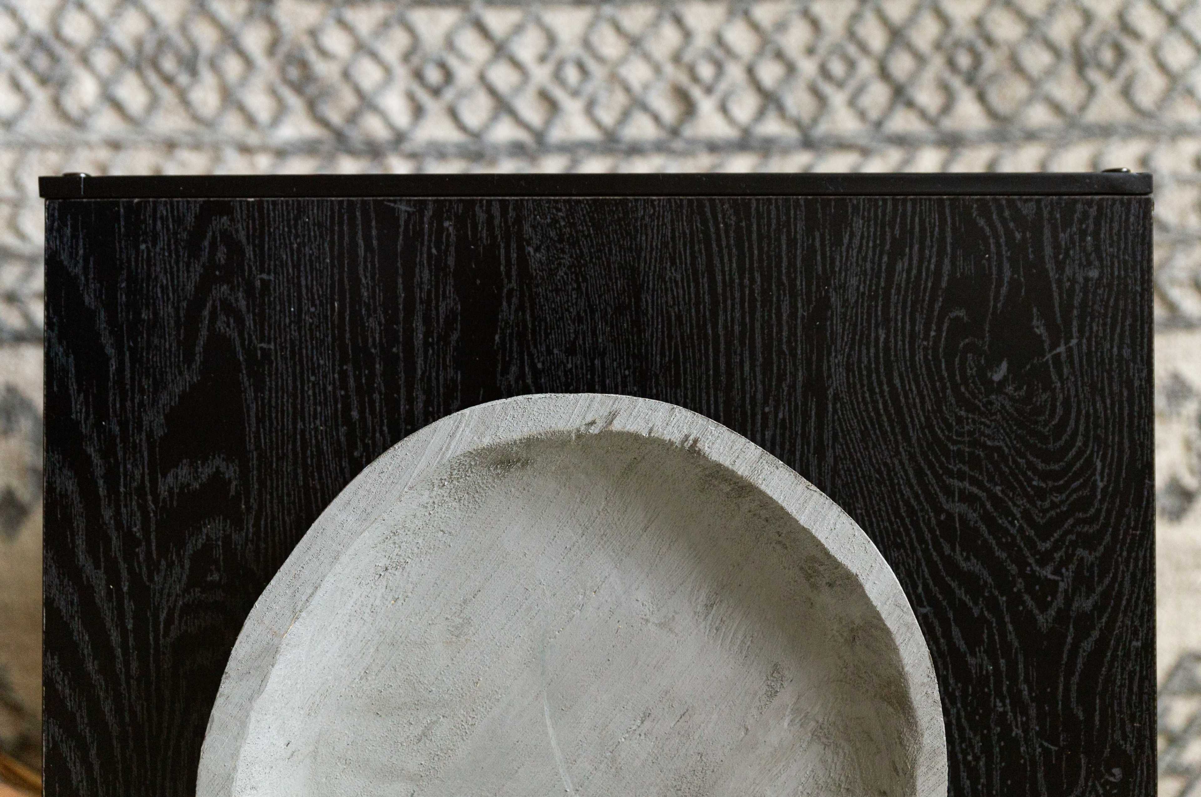 A shallow, round concrete bowl is placed on a dark wooden surface, with a patterned rug visible in the background.