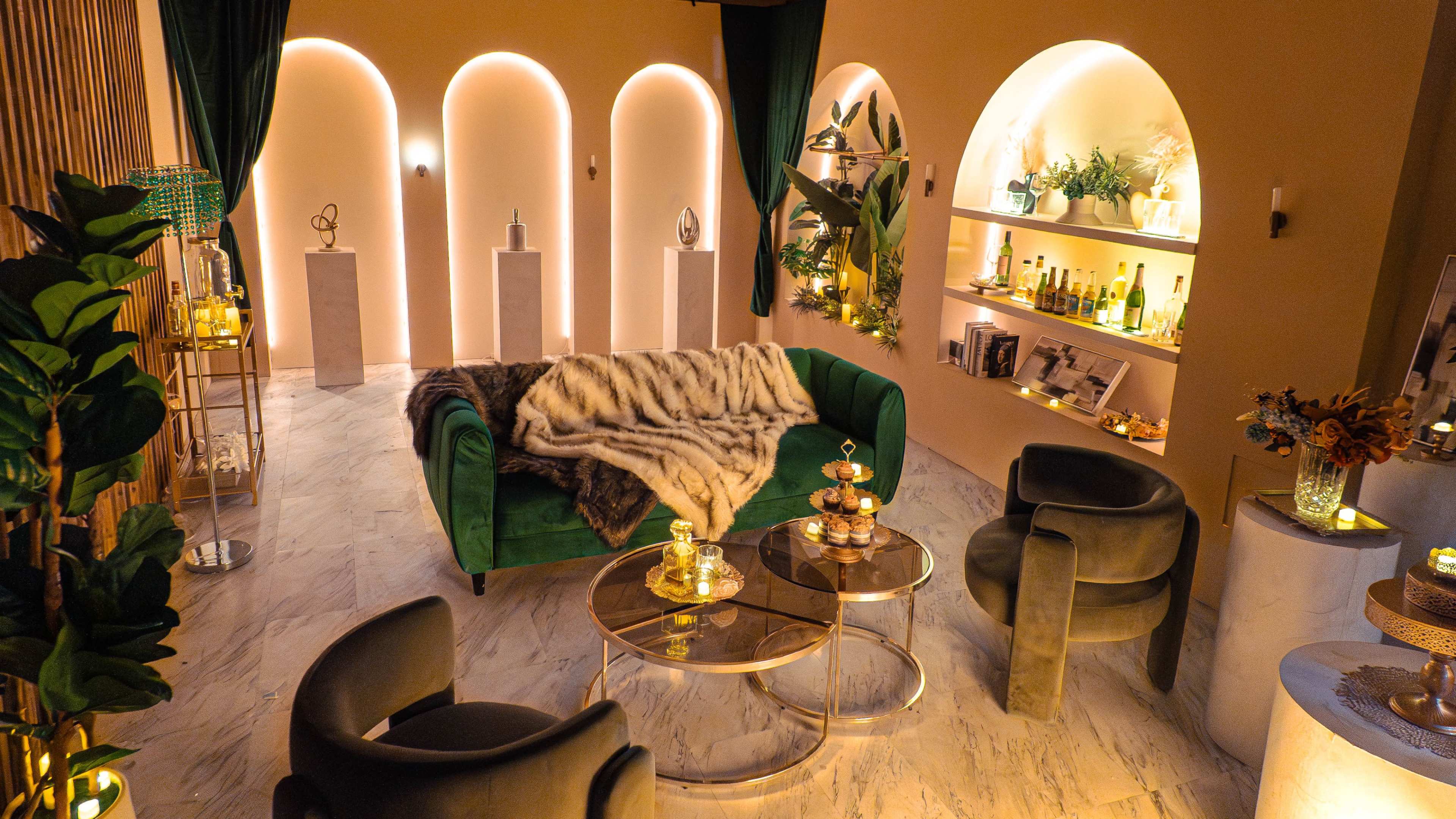 The image shows a modern lounge area with a green sofa, two black chairs, a coffee table, and shelves displaying decorative items and bottles.