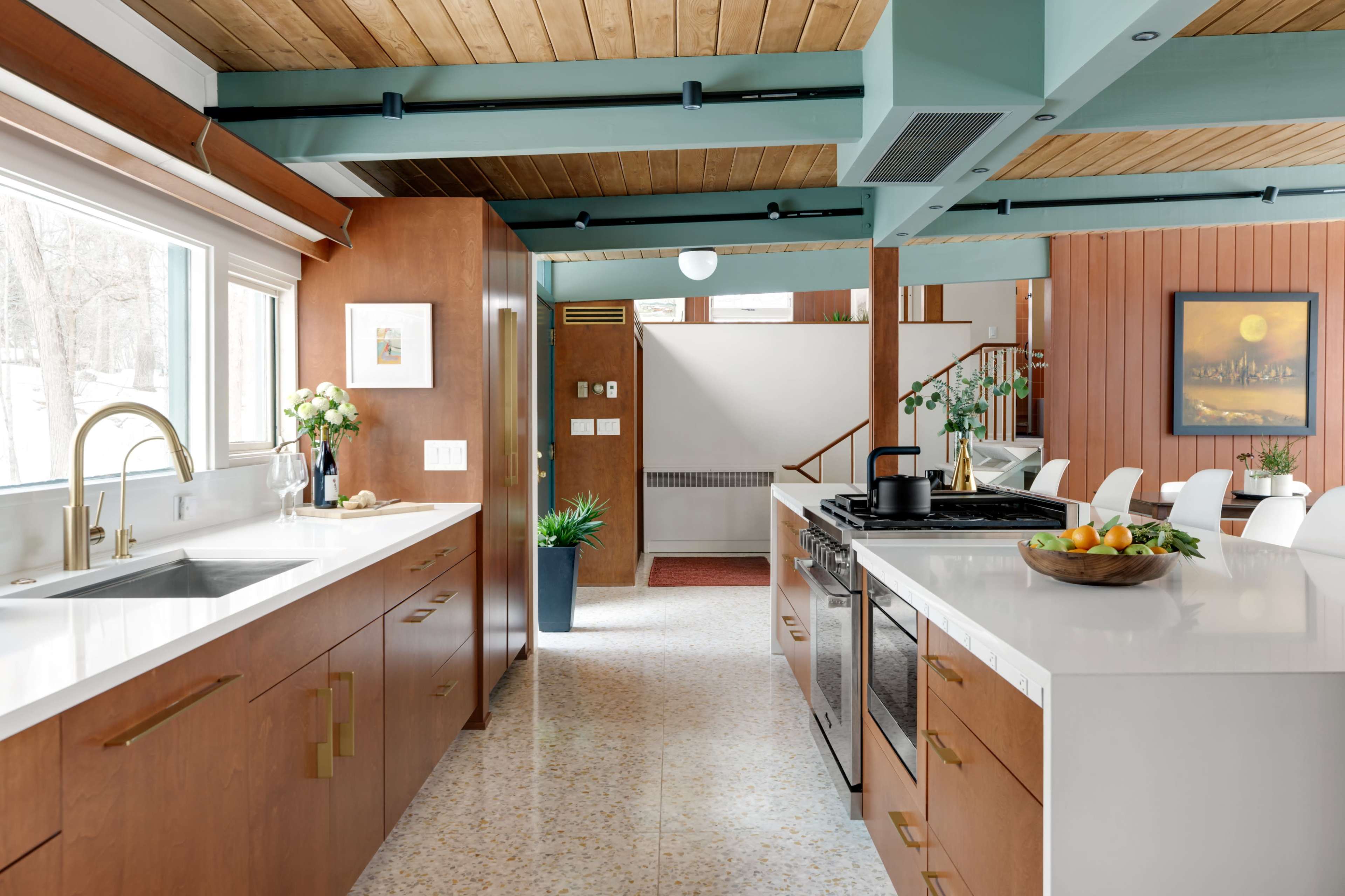 The image shows a modern kitchen with wooden cabinetry, white countertops, and a view into a dining area featuring a staircase and colorful decor.